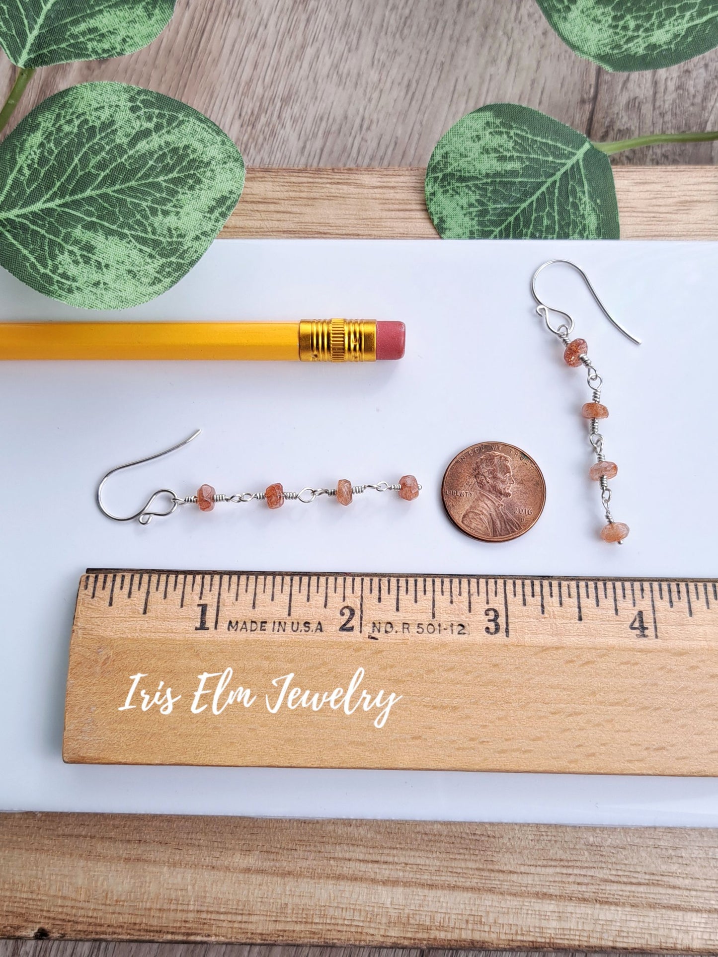 Two small beaded drop 2-3/8 inch long earrings with silver hooks and peach-orange beads are displayed on a white sheet with a wooden ruler, pencil, penny, and green leaves. The wooden ruler is branded 'Iris Elm Jewelry'.