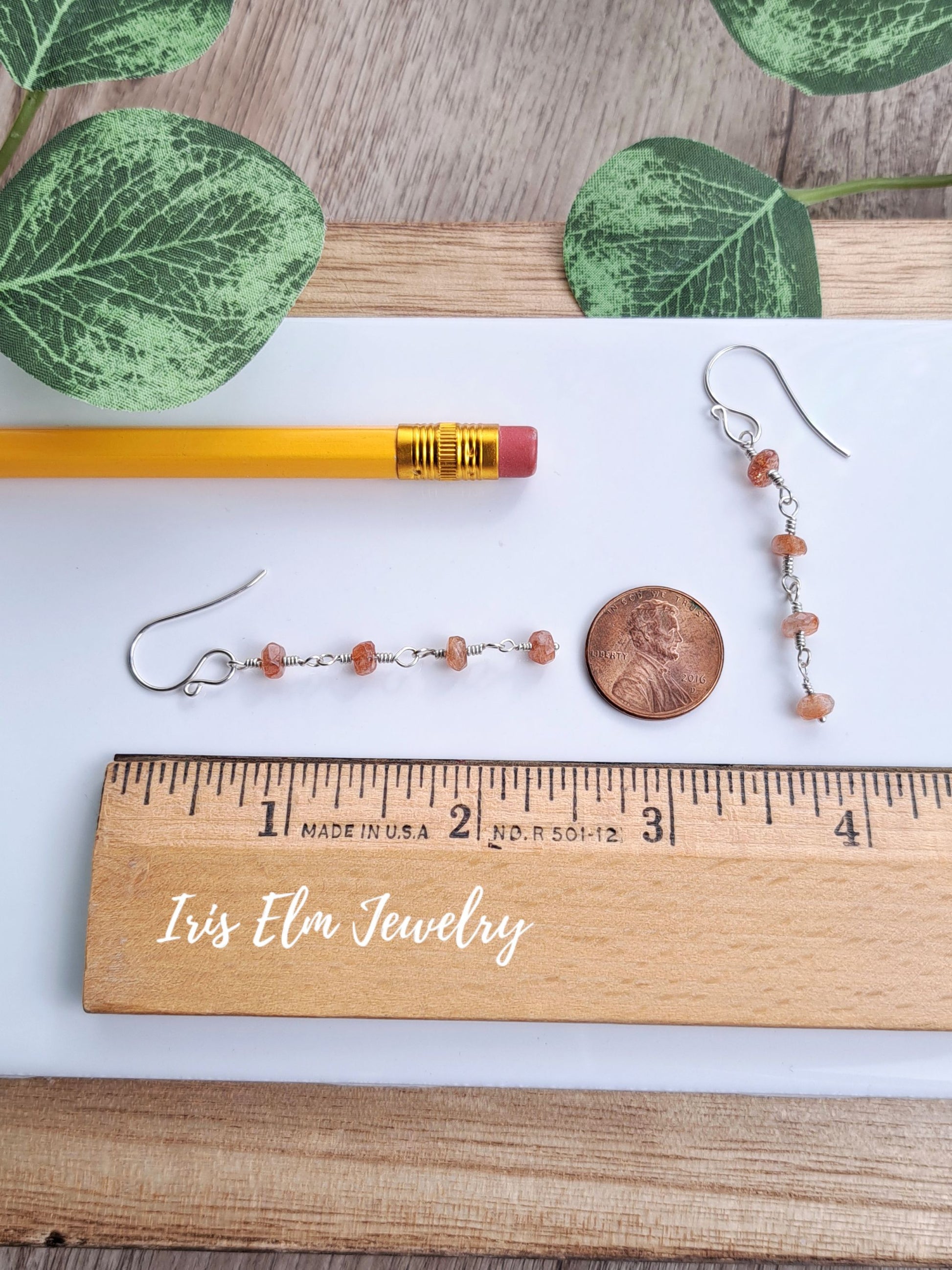 Two small beaded drop 2-3/8 inch long earrings with silver hooks and peach-orange beads are displayed on a white sheet with a wooden ruler, pencil, penny, and green leaves. The wooden ruler is branded 'Iris Elm Jewelry'.