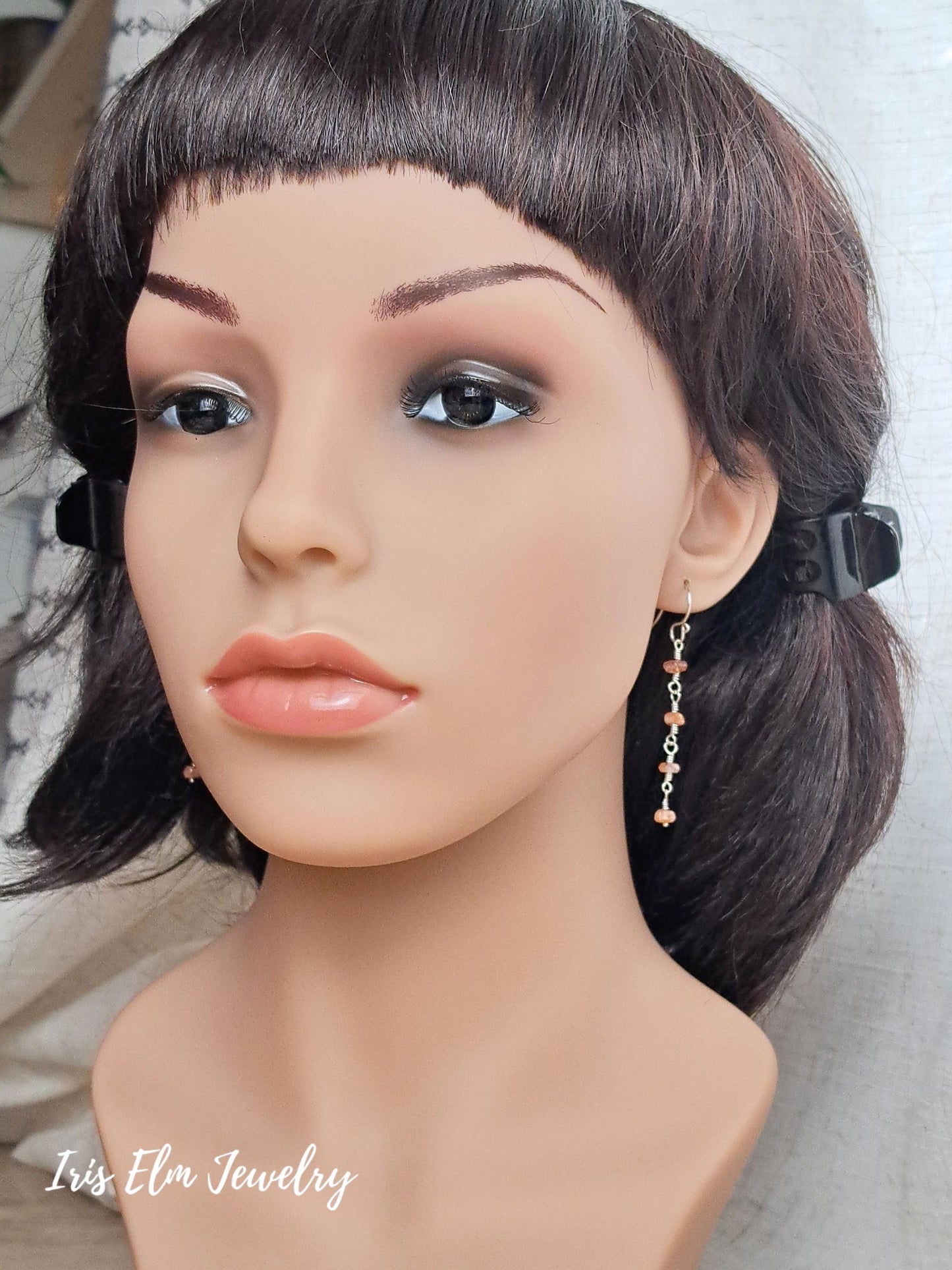 A mannequin bust wearing a short dark brown wig with bangs and dangling gold-toned earrings with small pink and orange bead accents. The mannequin is photographed against a light neutral background.