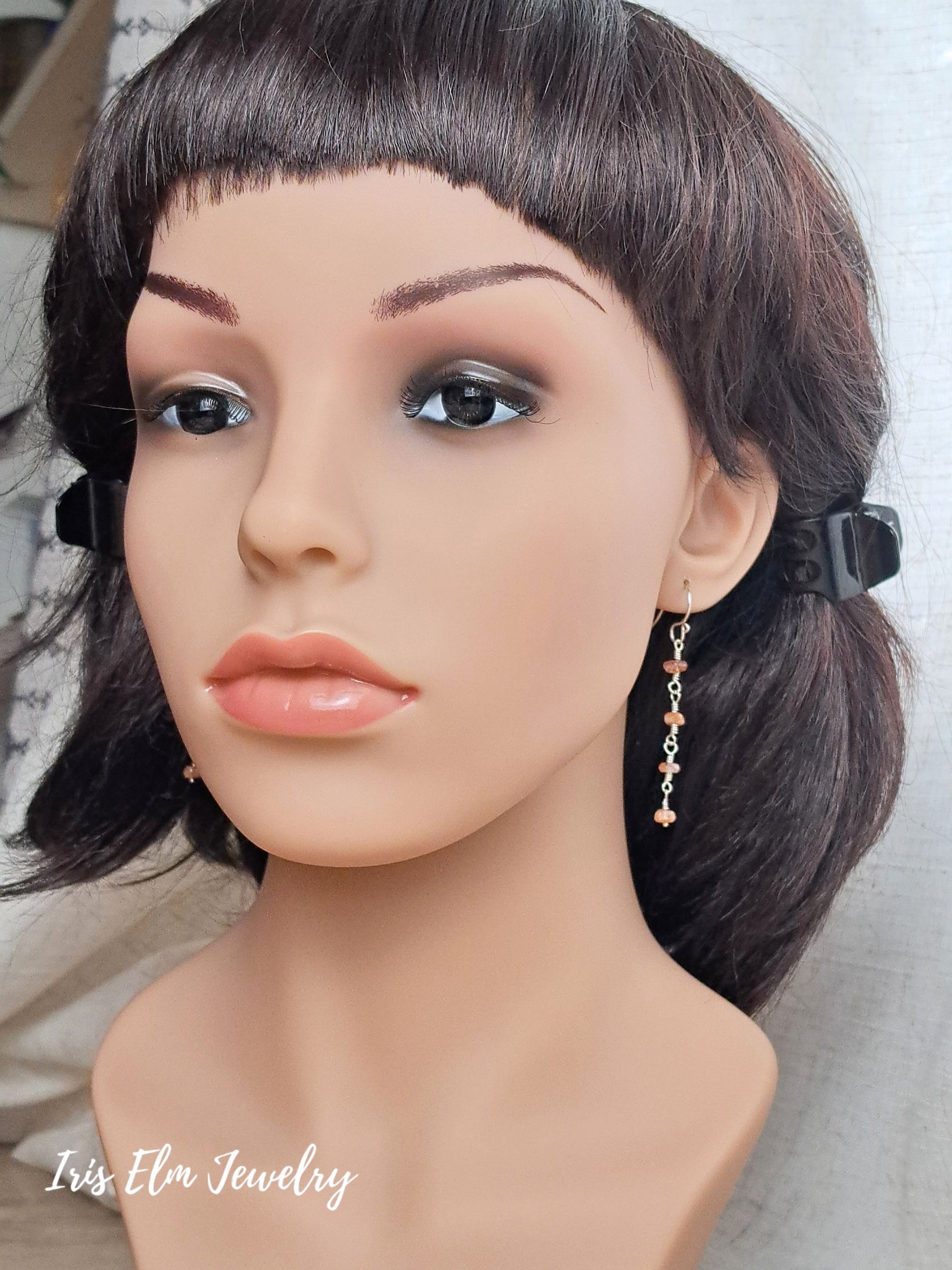A mannequin bust wearing a short dark brown wig with bangs and dangling gold-toned earrings with small pink and orange bead accents. The mannequin is photographed against a light neutral background.