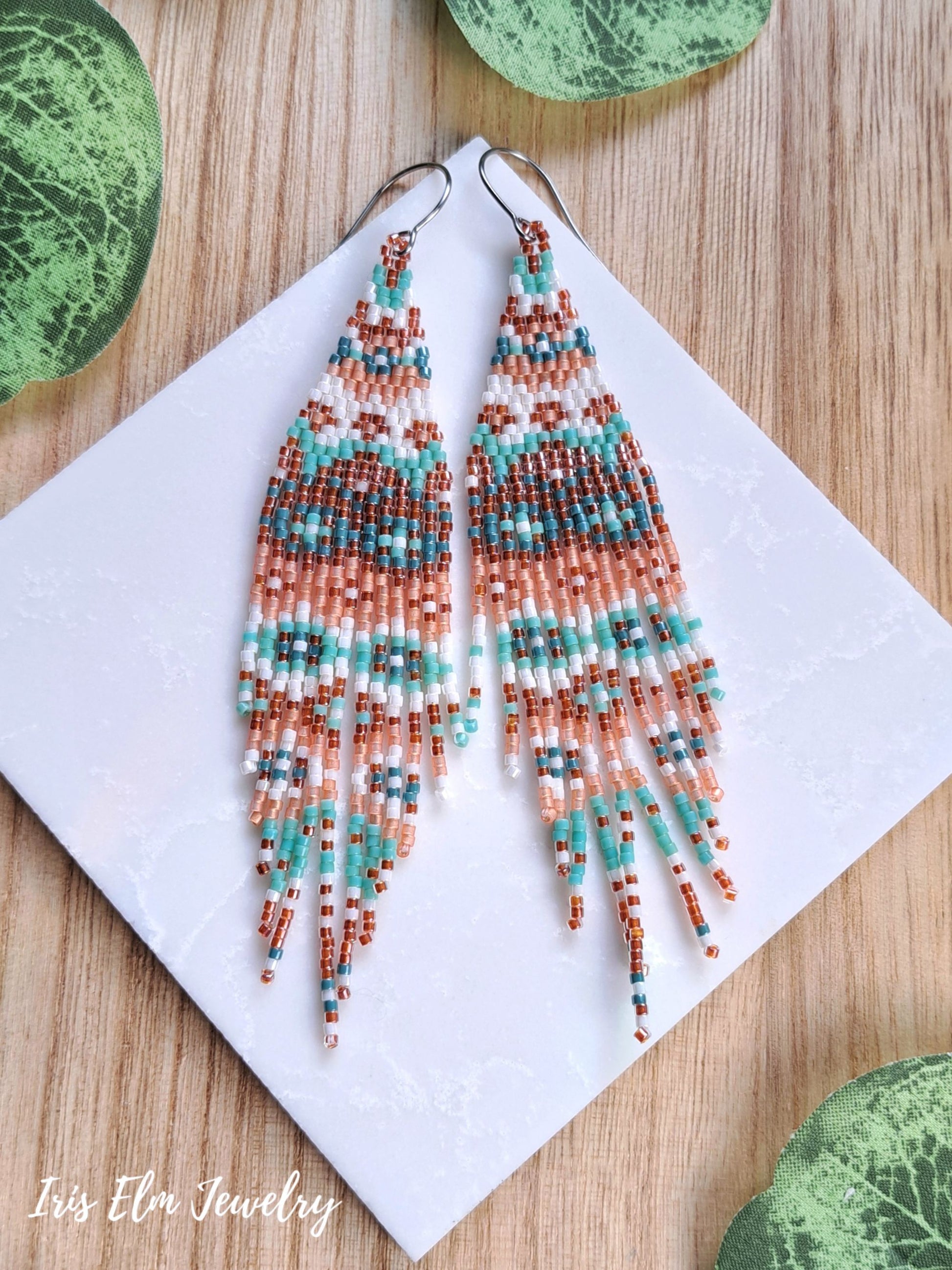 A pair of long beaded dangle earrings with a geometric pattern in turquoise, rust orange, white, and mint, displayed on a white square card over a wooden surface with green leaves.
