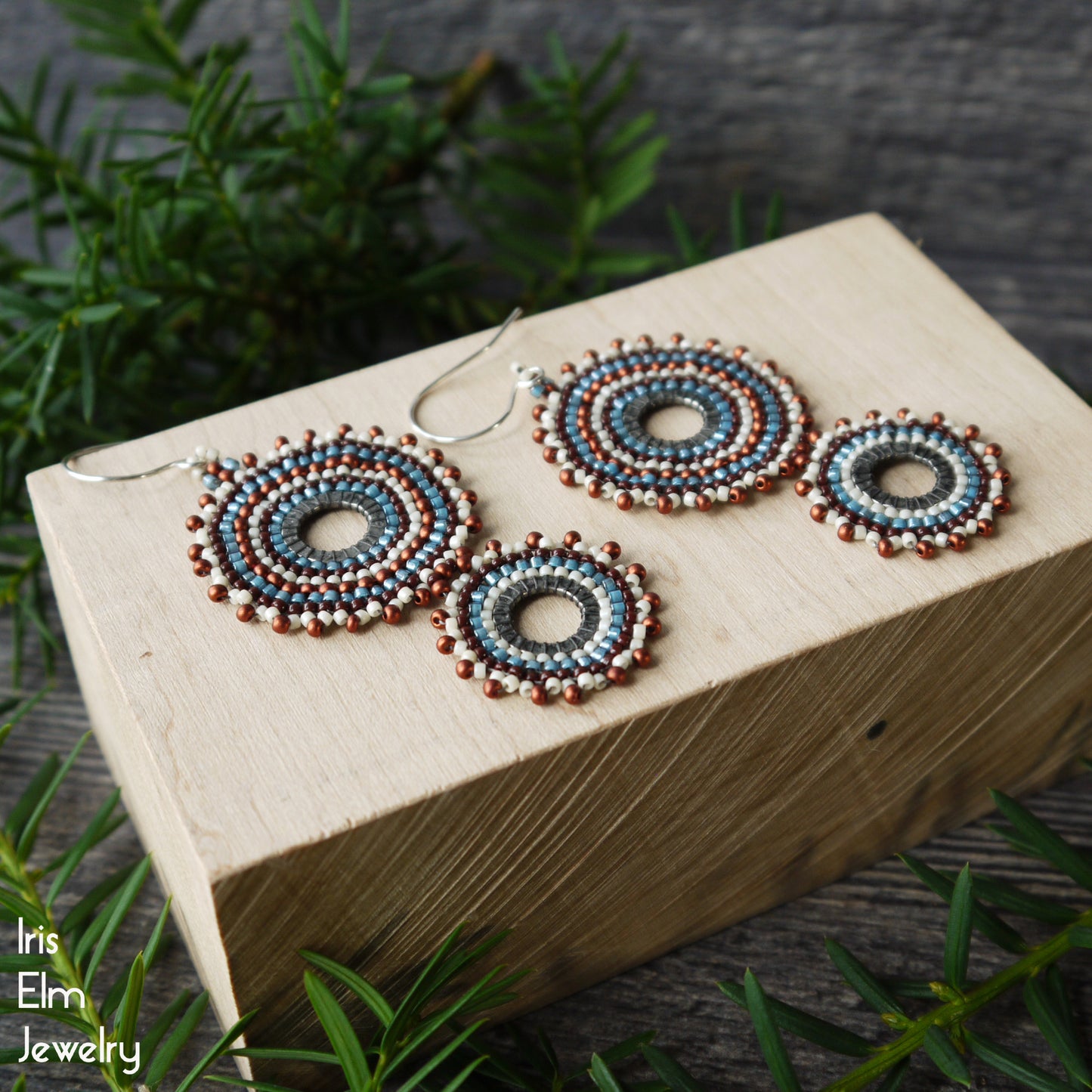 Seed bead beaded hoop earrings displayed on a light wood block with green foliage in the background. The earrings feature concentric rings of blue, white, and copper-brown seed beads with sterling silver fishhook wires.