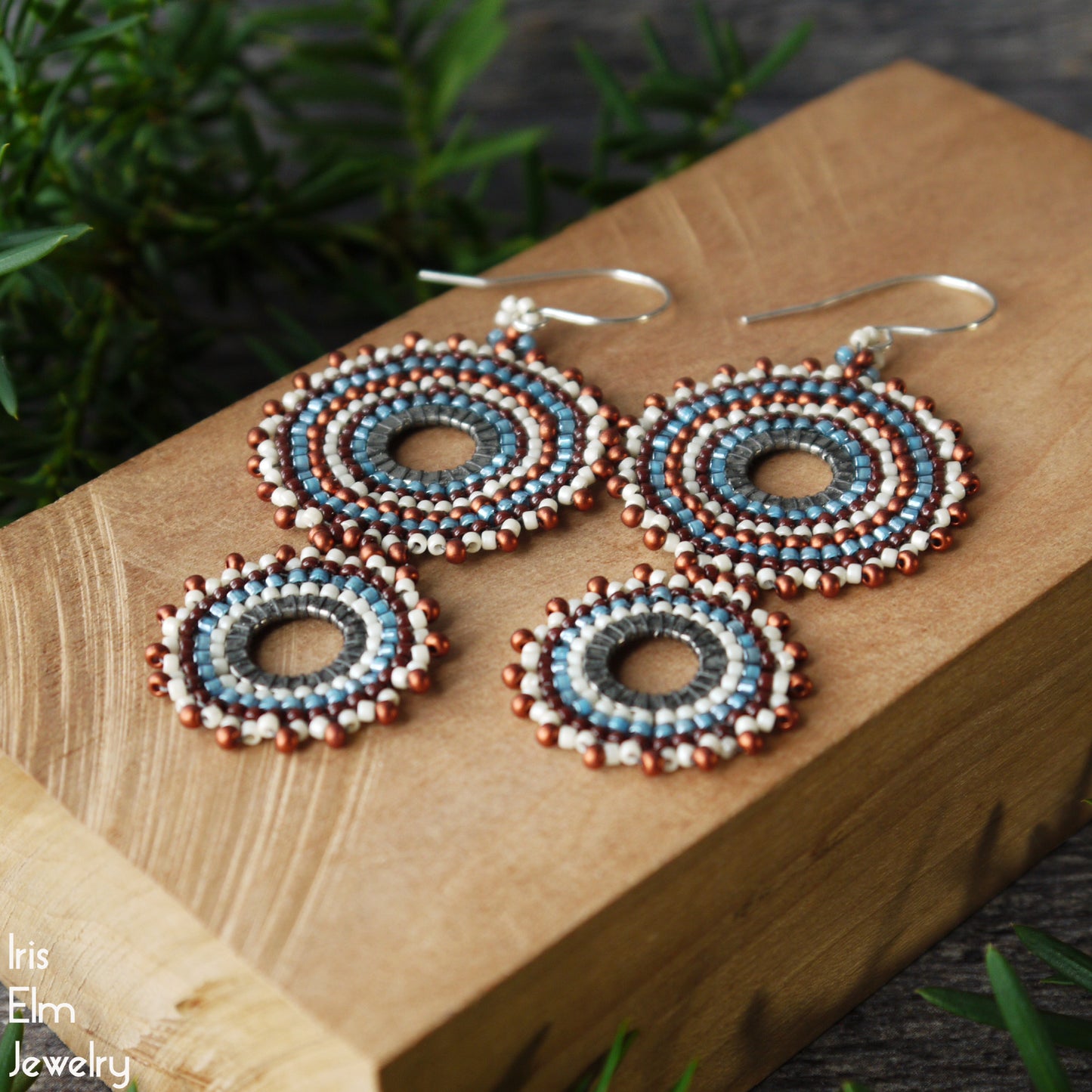 Handmade beaded dangle earrings with circular concentric patterns in blue, white, and reddish-brown, displayed on a wooden block with green foliage in the background.