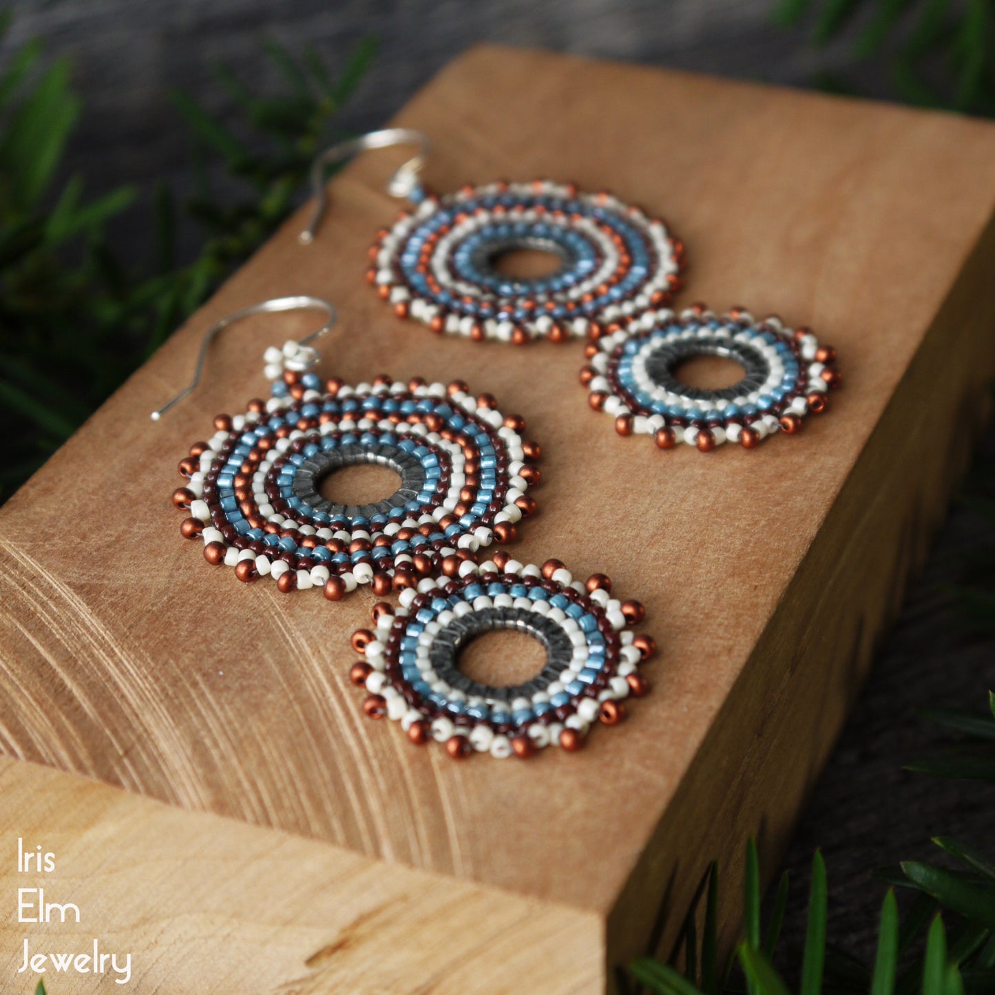 Handmade beaded hoop earrings with blue, white, and copper-red concentric circles, displayed on a wooden block with the brand name 'Iris Elm Jewelry' in the bottom left corner.