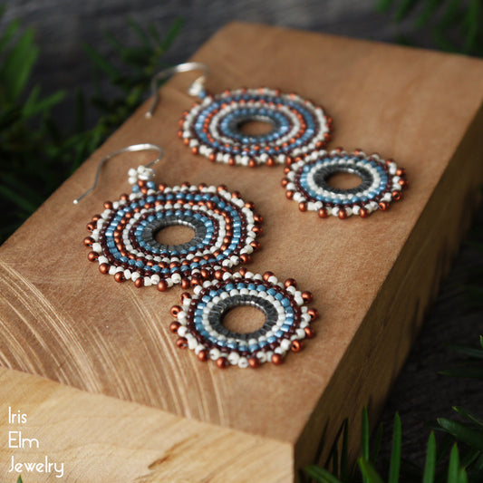 Handmade beaded hoop earrings with blue, white, and copper-red concentric circles, displayed on a wooden block with the brand name 'Iris Elm Jewelry' in the bottom left corner.