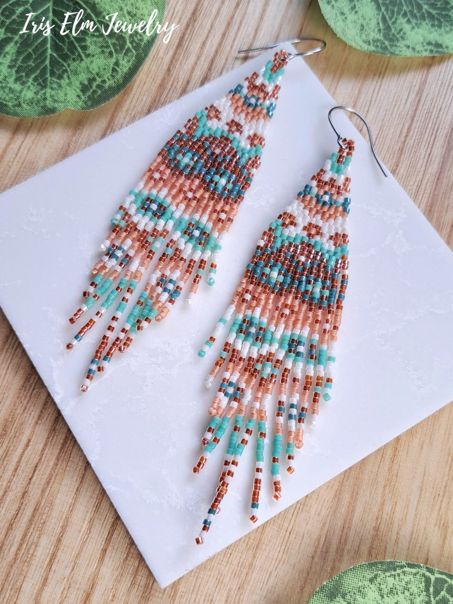 A pair of long beaded fringe earrings displayed on a white square card over a light wood surface, with green leaves partially visible around the edges. The brand name 'Iris Elm Jewelry' appears in the top left corner.