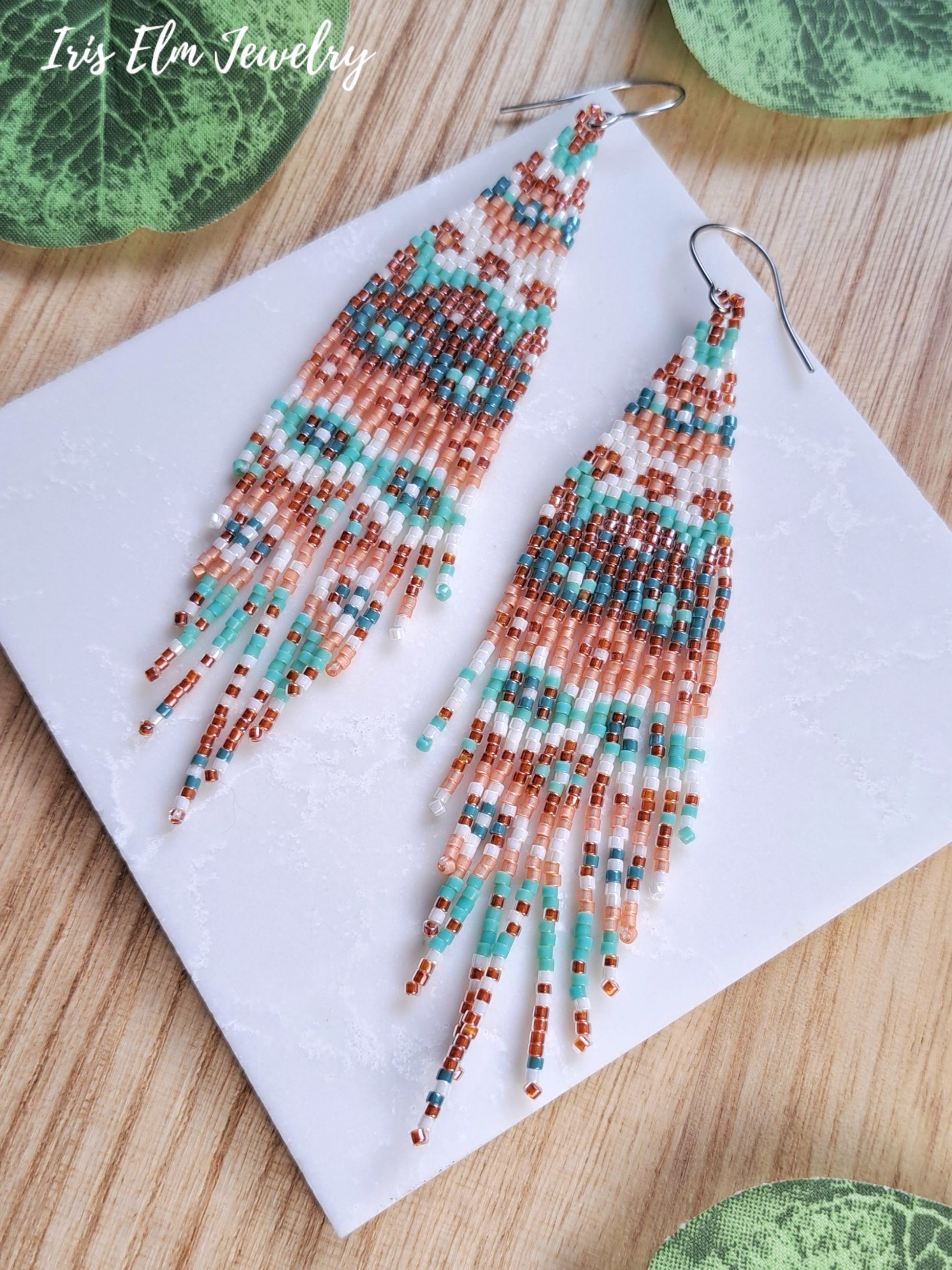 A pair of long beaded fringe earrings displayed on a white square card over a light wood surface, with green leaves partially visible around the edges. The brand name 'Iris Elm Jewelry' appears in the top left corner.