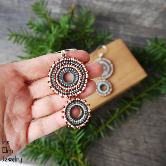 A hand holds a pair of handmade beaded dangle earrings with circular concentric patterns in blue, gray, and rose gold. The earrings are shown against a rustic background with pine branches and a wooden block, with the brand name 'Iris Elm Jewelry' visible in the bottom left corner.