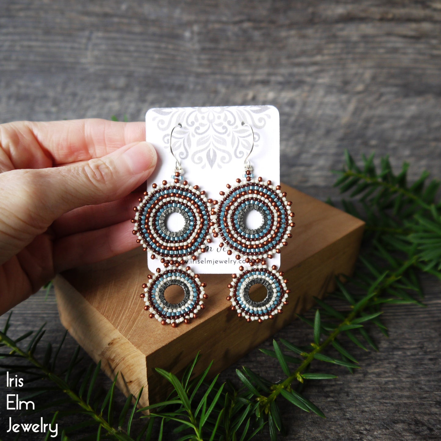 A hand holds a display card showing a pair of handmade beaded earrings with circular patterns in teal, brown, and white, set on a wooden block with pine branches.