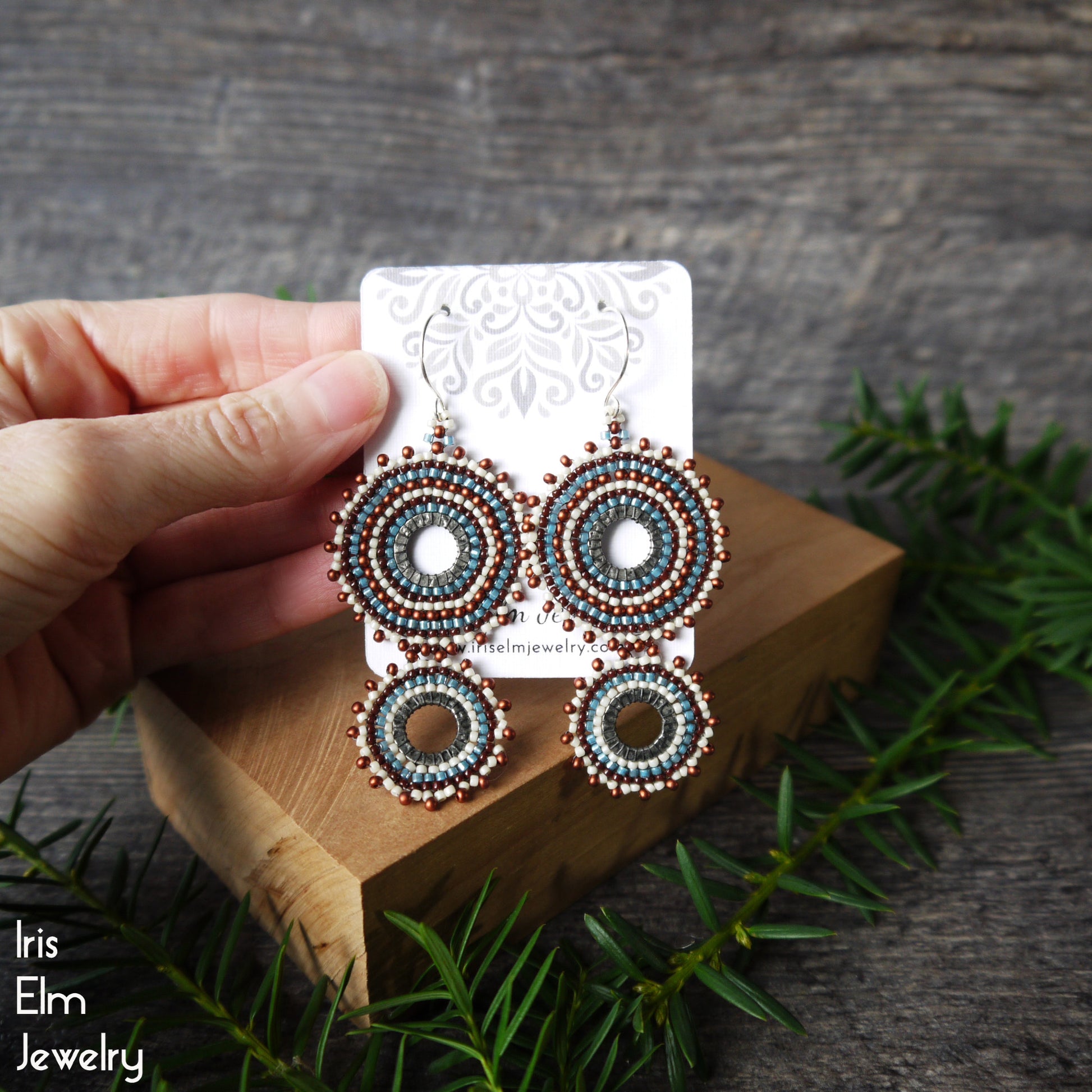 A hand holds a display card showing a pair of handmade beaded earrings with circular patterns in teal, brown, and white, set on a wooden block with pine branches.