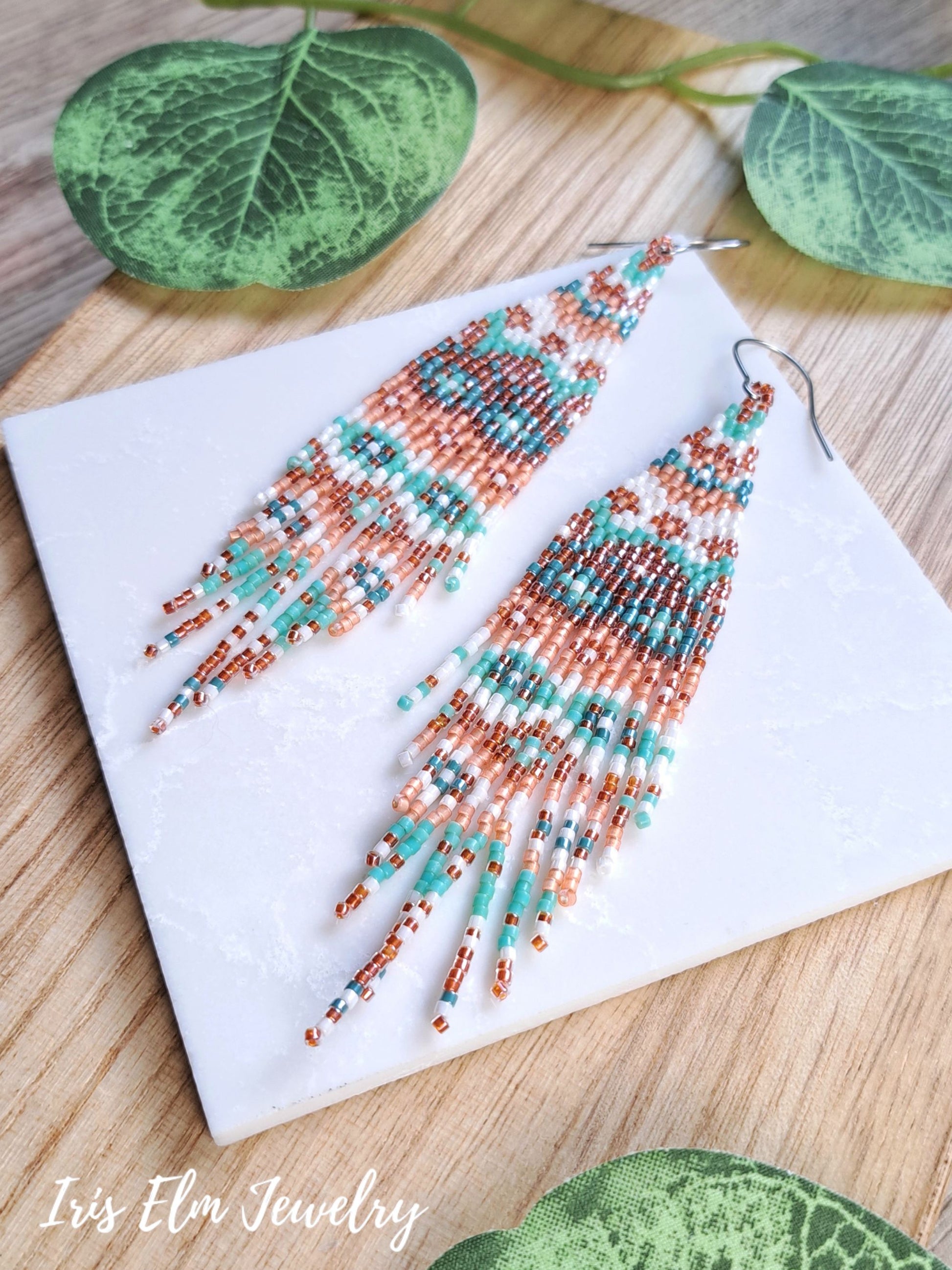 A pair of long beaded fringe earrings displayed on a white square card over a wooden surface with decorative green leaves. The earrings feature a geometric pattern in turquoise, copper, white, and dark blue seed beads with silver fishhook wires.