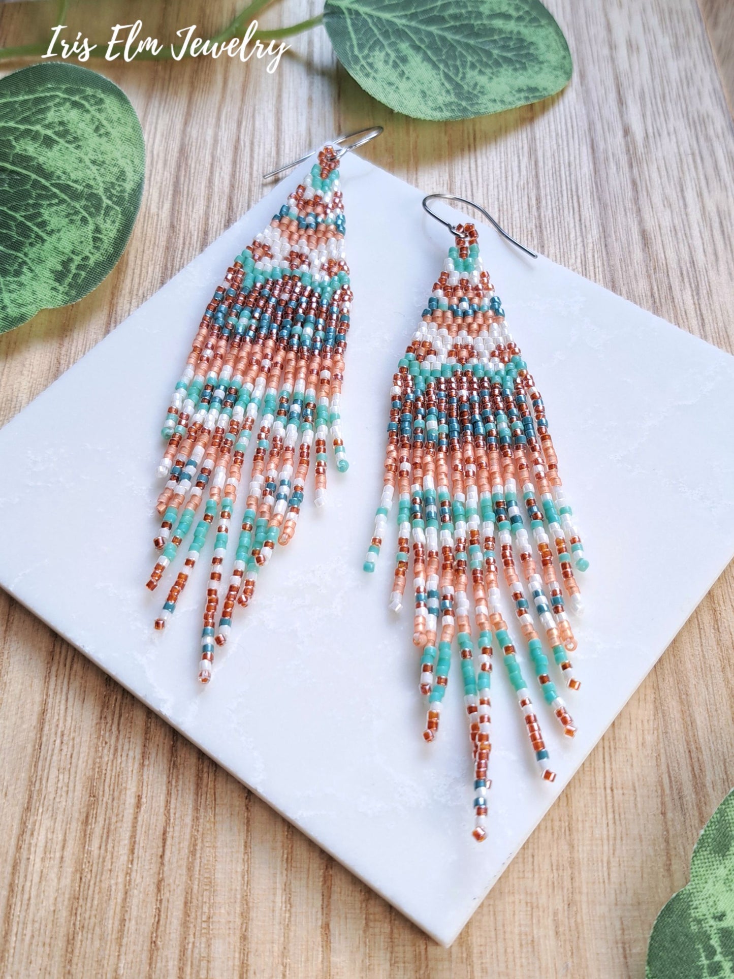A pair of long beaded fringe earrings displayed on a white square tile over a wood surface, with green leaves around the edges. The earrings feature geometric bead patterns in turquoise, rust orange, white, and mint green.