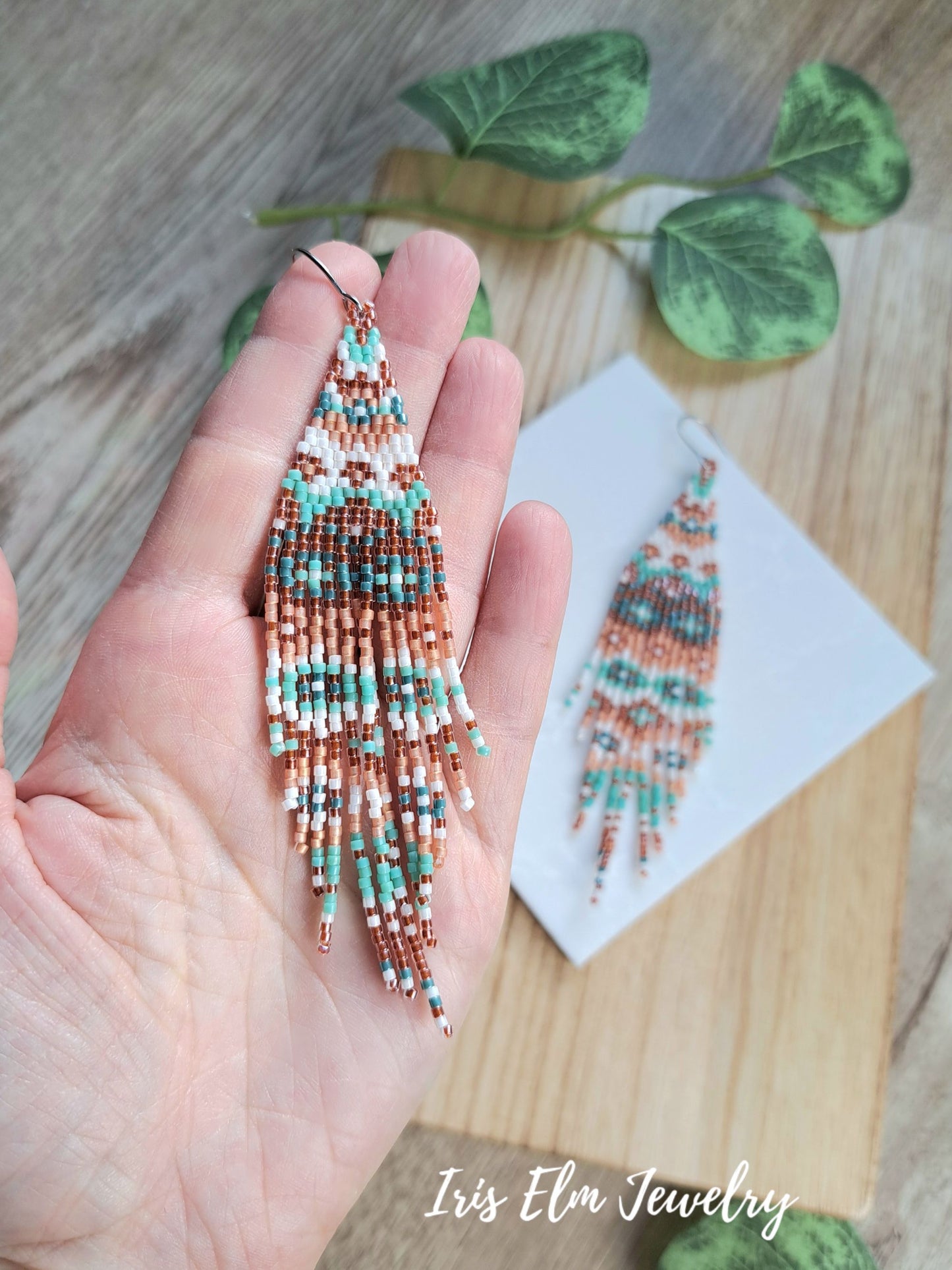 A hand holds a pair of long beaded fringe earrings with a geometric pattern in turquoise, white, and copper-peach. A second matching earring is displayed on a small white card on a wooden surface with green leaves in the background.