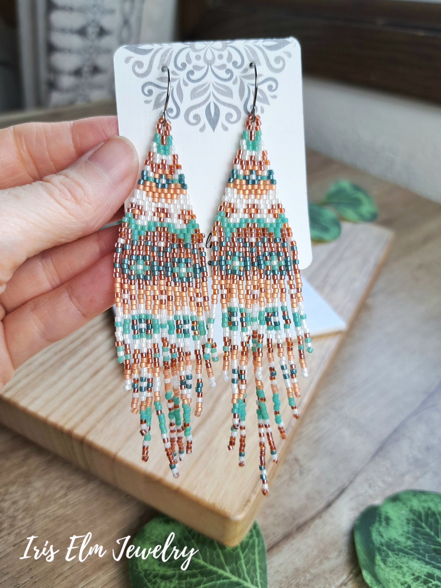 A hand holds a pair of long beaded fringe earrings on a white display card. The earrings feature intricate geometric beadwork in turquoise, white, and copper-peach, hanging from silver-toned hooks.