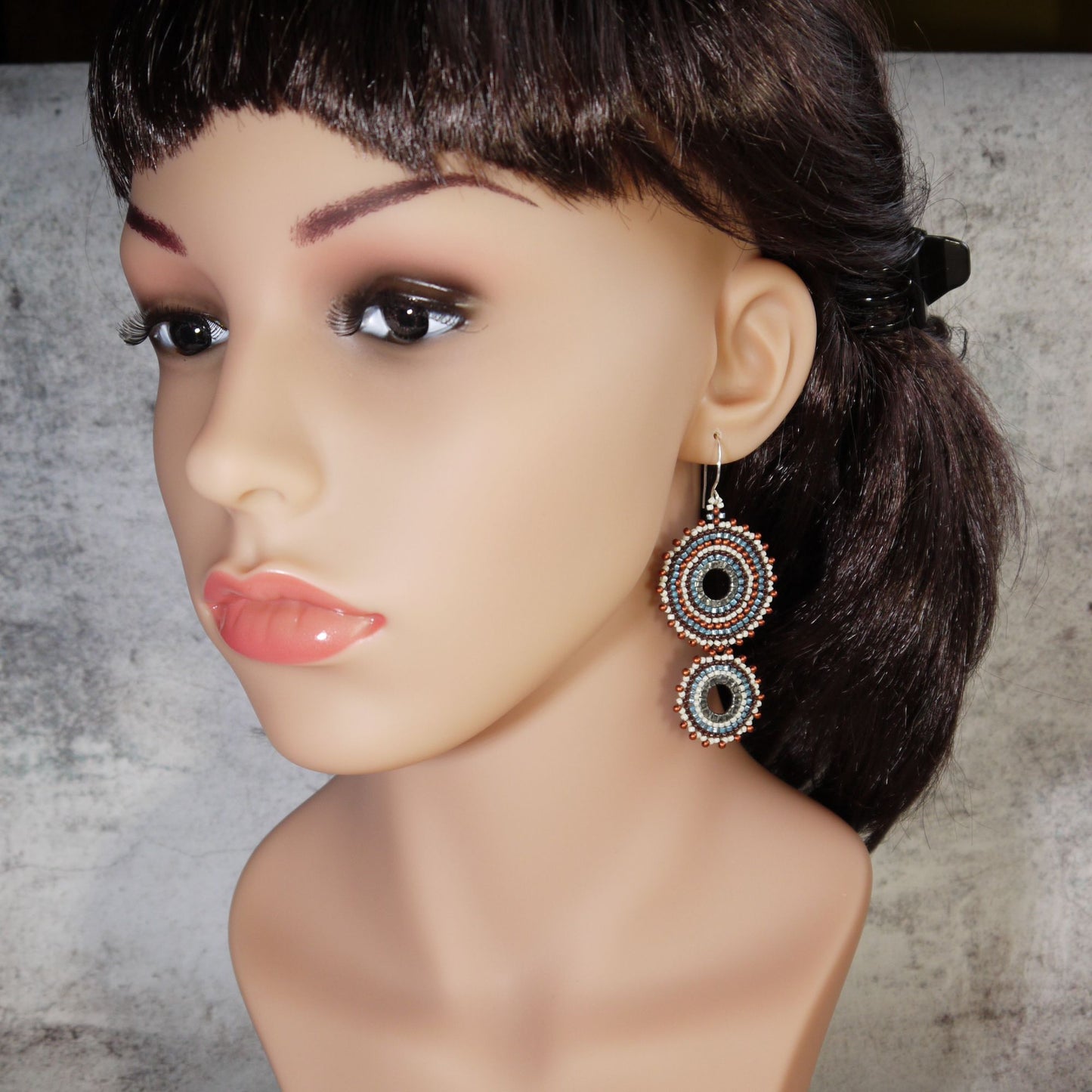 A pair of dangling circular earrings displayed on a mannequin bust. The earrings feature concentric rings in black, white, and metallic silver tones.