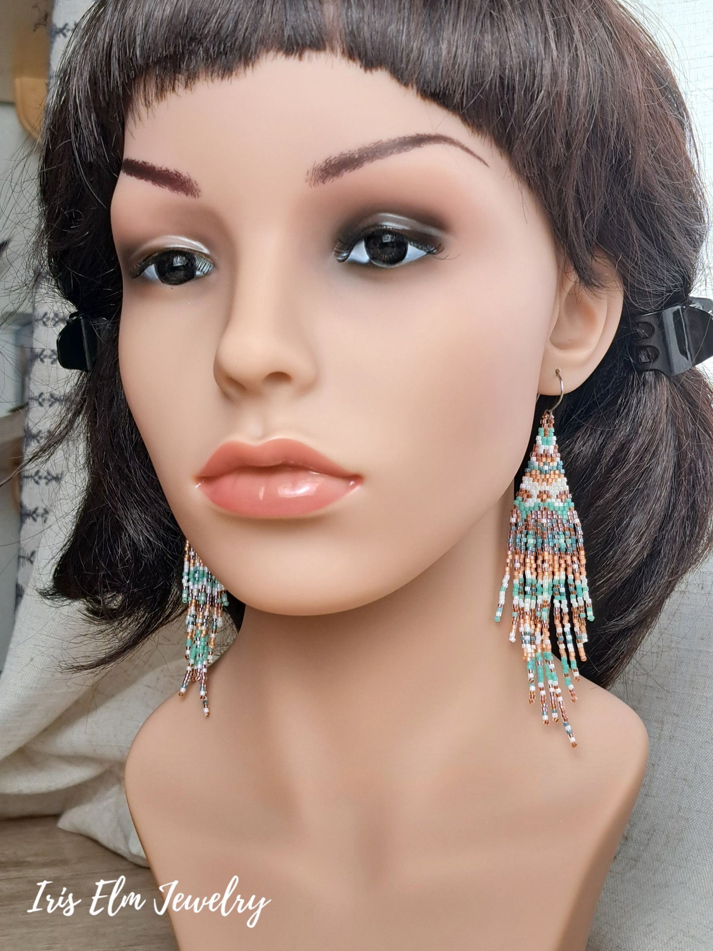 A mannequin bust wearing a short dark wig and large dangling beaded fringe earrings. The earrings have turquoise, coral, pink, and mint beads.