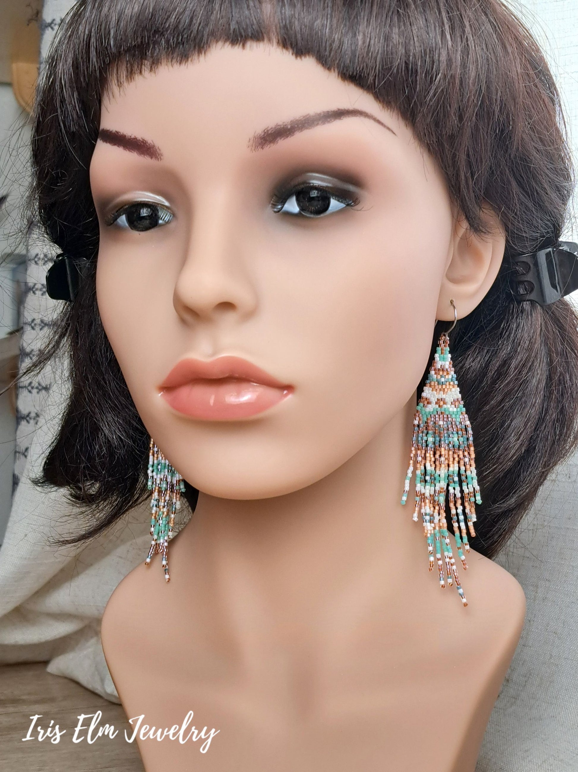 A mannequin bust wearing a short dark wig and large dangling beaded fringe earrings. The earrings have turquoise, coral, pink, and mint beads.