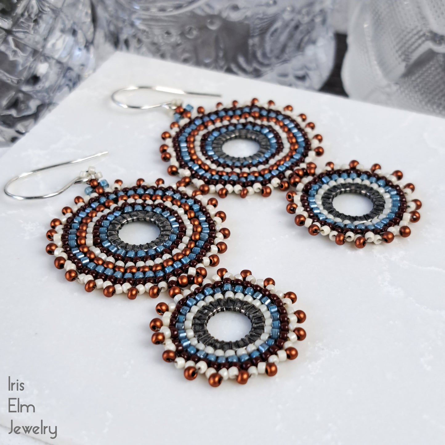 Three circular beaded dangle earrings with silver fishhook wires, featuring concentric rings of blue, brown, white, and copper-brown beads. The earrings are displayed on a white textured surface with a blurred background.