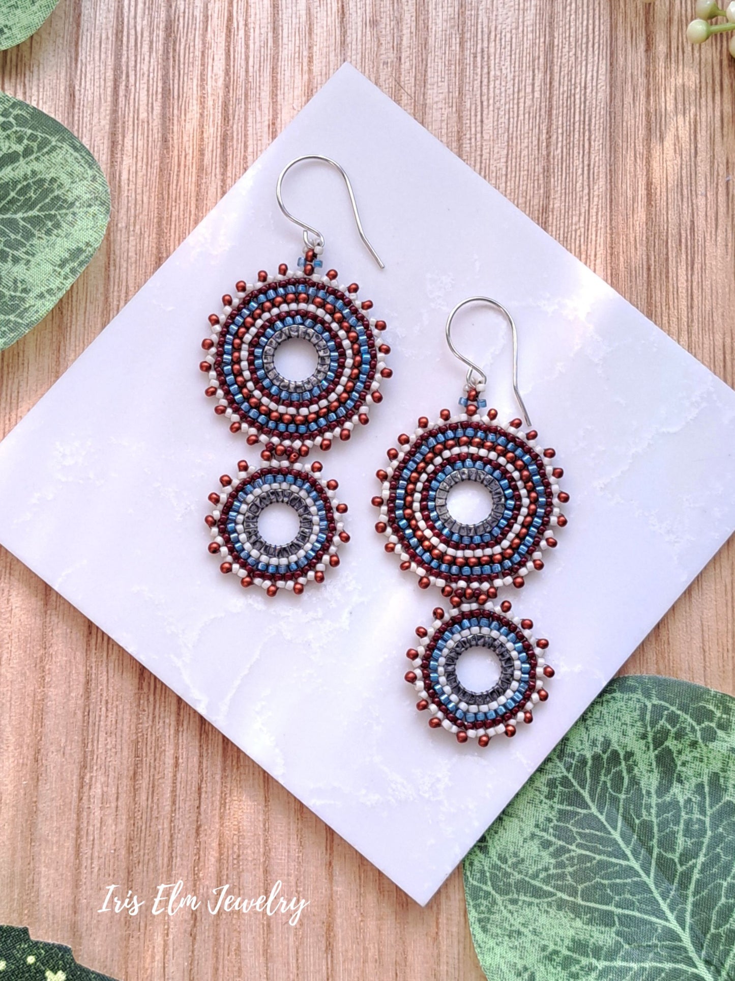 A pair of handmade beaded dangle earrings displayed on a white square card, set against a light wood background with decorative green leaves. The earrings feature intricate circular beadwork in shades of blue, burgundy, and burnt sienna, suspended from silver fishhook wires.