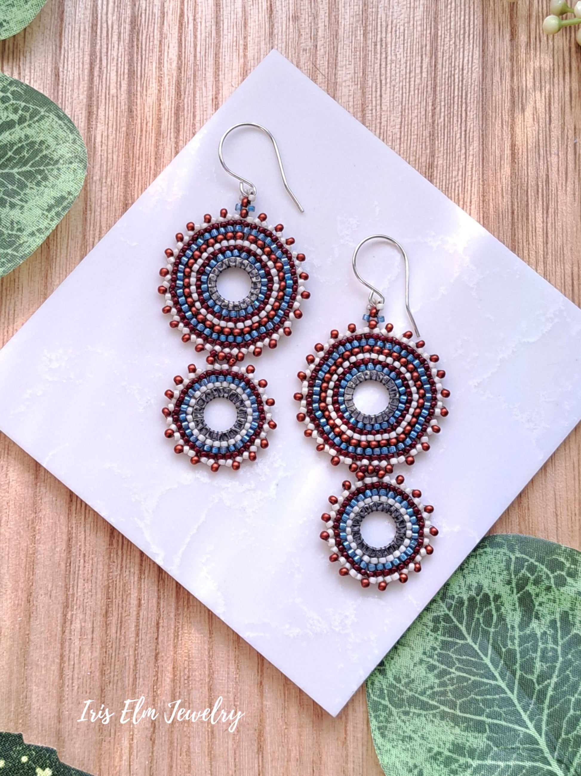 A pair of handmade beaded dangle earrings displayed on a white square card, set against a light wood background with decorative green leaves. The earrings feature intricate circular beadwork in shades of blue, burgundy, and burnt sienna, suspended from silver fishhook wires.