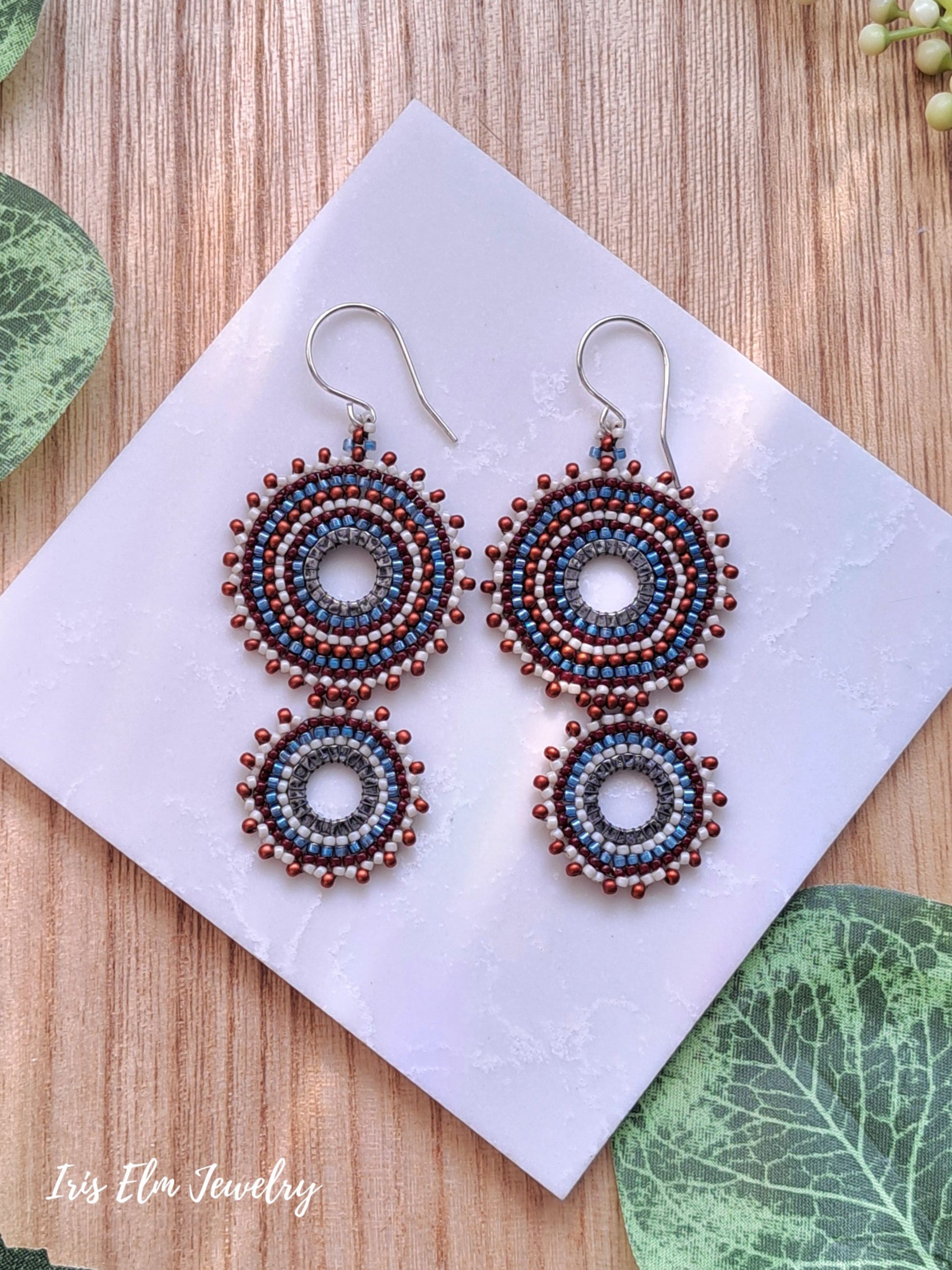 A pair of handmade beaded hoop seed bead dangle earrings with circular spiral patterns in blue, cream, and reddish-brown, displayed on a white card over a wooden surface with decorative green leaves.