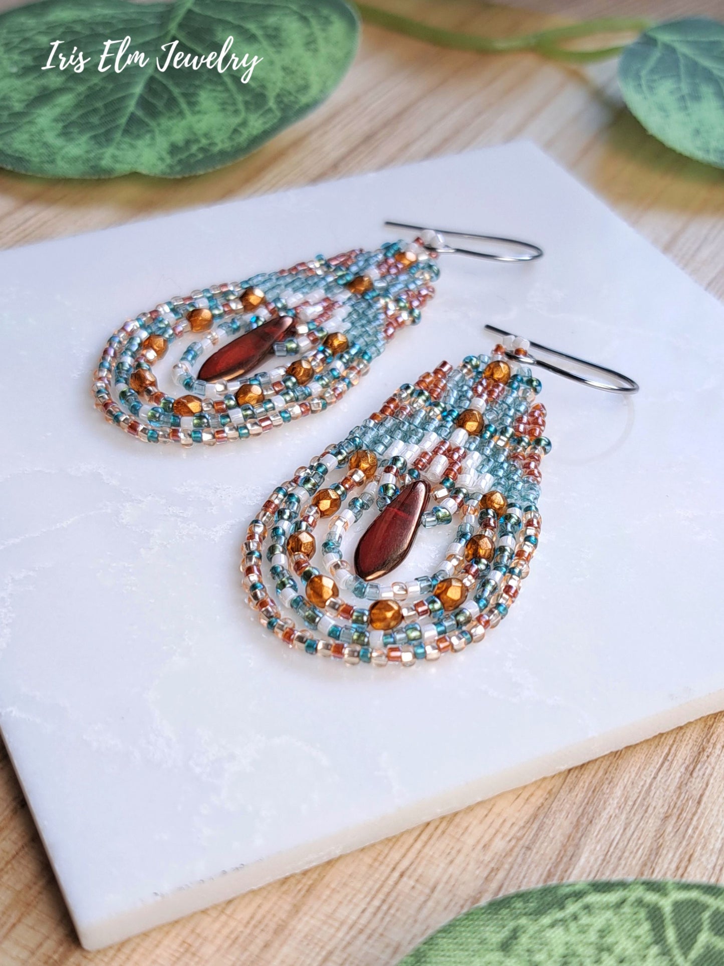 A pair of handmade beaded teardrop dangle earrings by Iris Elm Jewelry, featuring turquoise, copper, and white seed beads with a central burnt orange teardrop glass bead, displayed on a white tile.