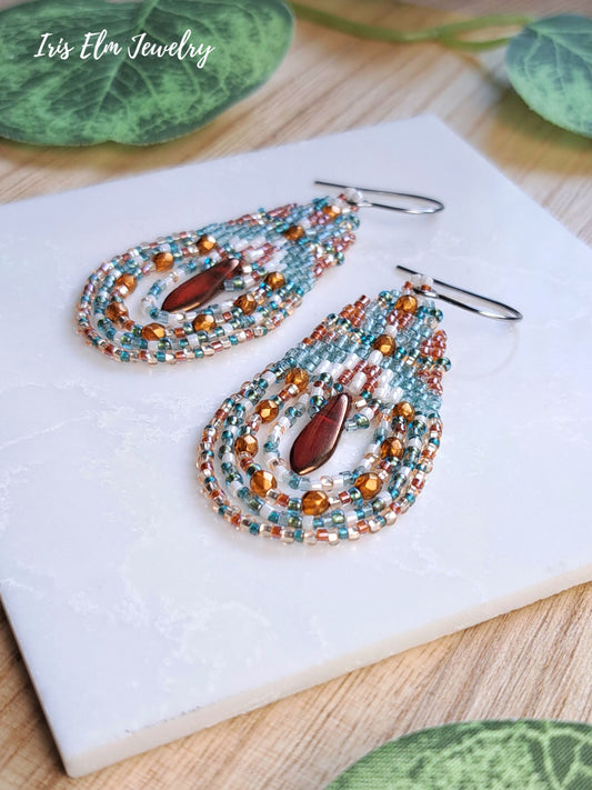 A pair of handmade beaded teardrop dangle earrings by Iris Elm Jewelry, featuring turquoise, copper, and white seed beads with a central burnt orange teardrop glass bead, displayed on a white tile.
