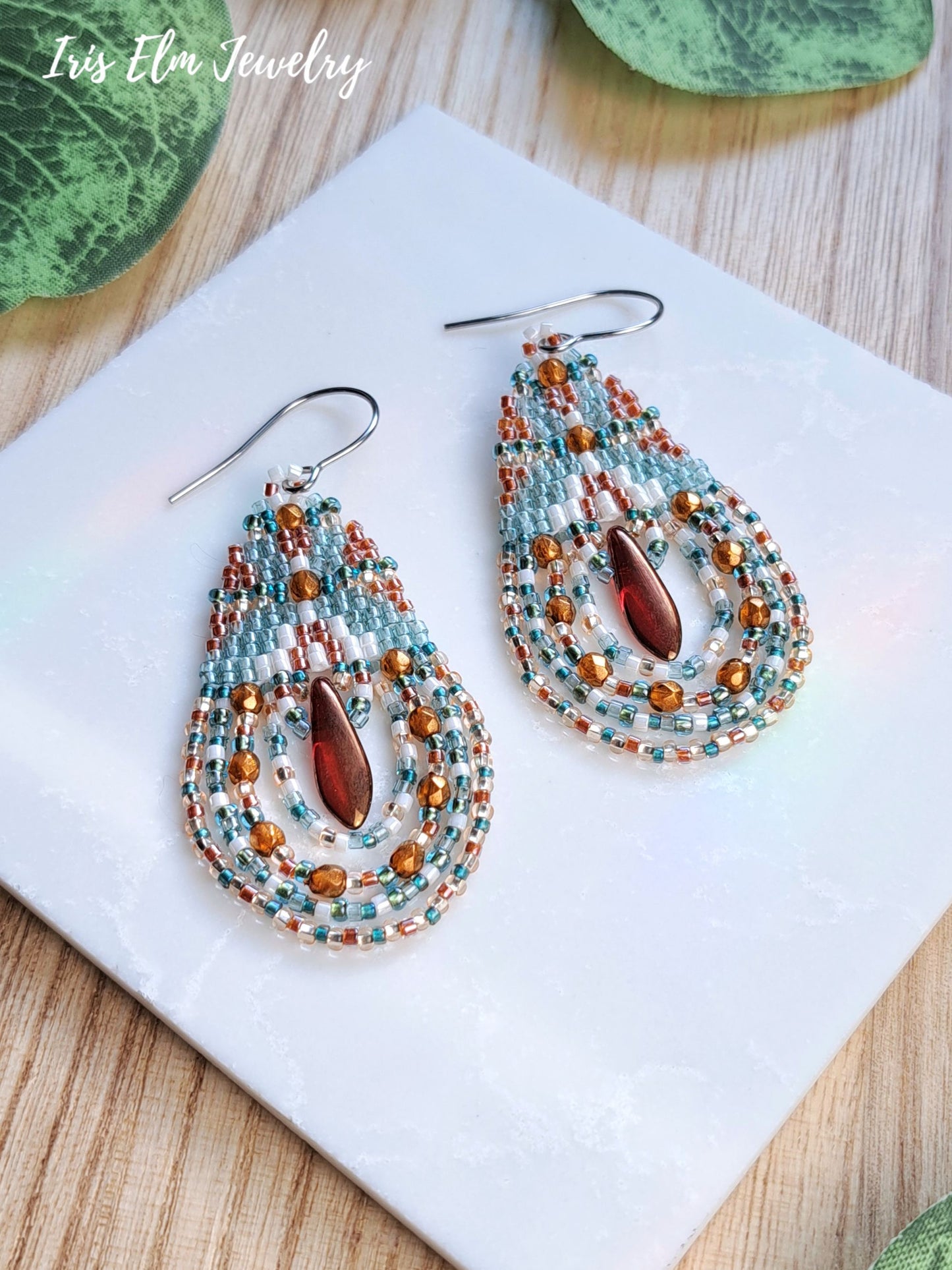 A pair of handmade teardrop-shaped beaded earrings by Iris Elm Jewelry, featuring turquoise, white, and copper seed beads with a central amber teardrop glass bead, displayed on a white square tile over a wooden surface.