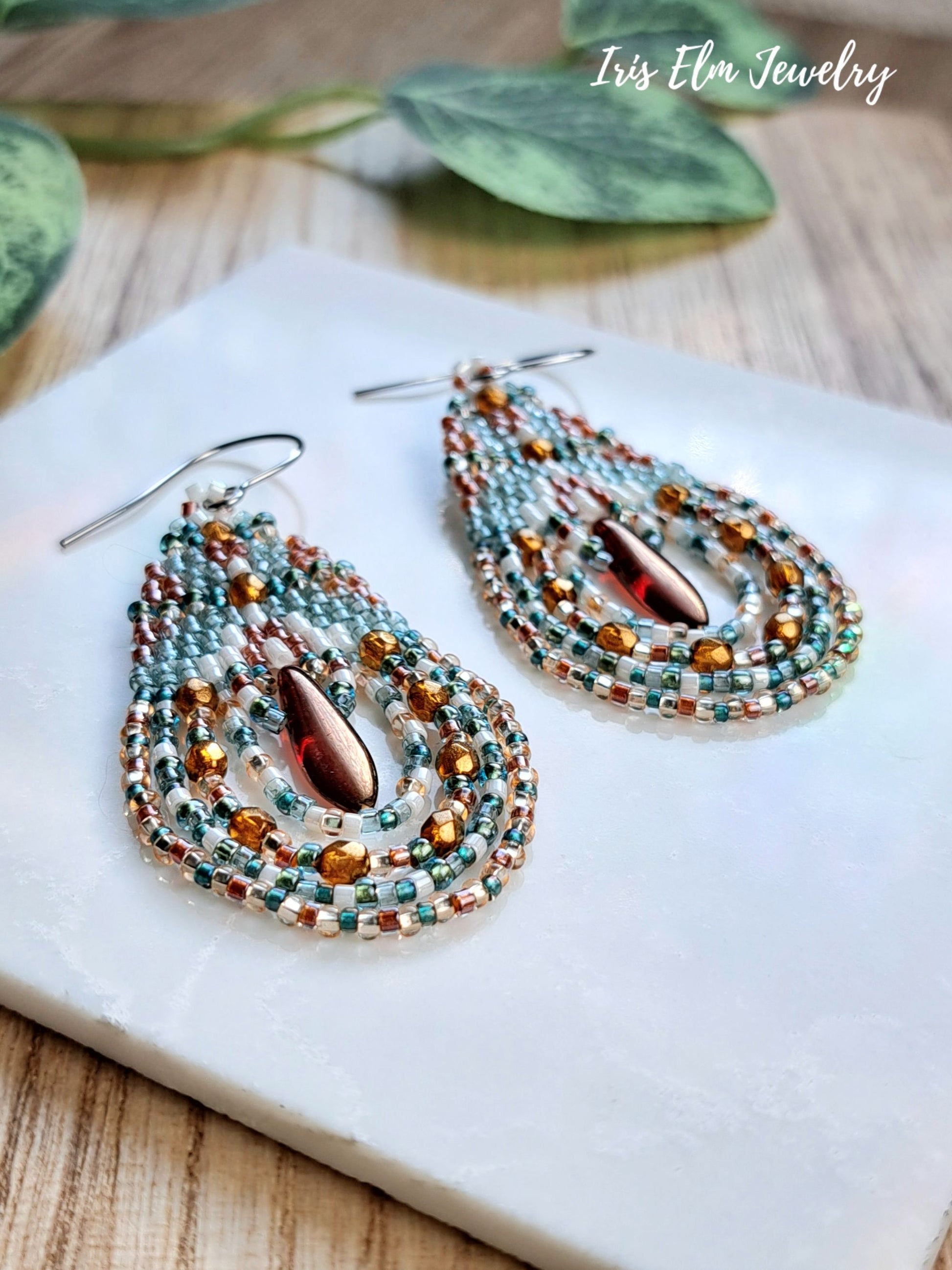 A pair of handmade beaded teardrop earrings with silver fishhook wires, featuring layered rows of teal, copper, and white seed beads surrounding a central amber-brown teardrop bead. The earrings are displayed on a white stone slab over a wooden surface with green leaves in the background.