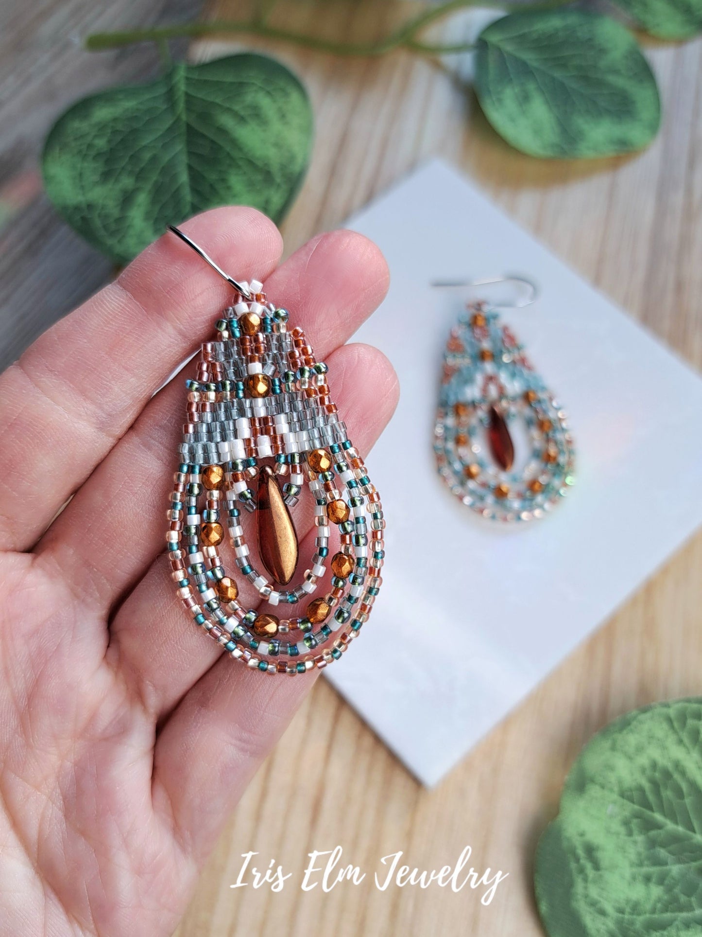 A hand holds a beaded teardrop earring in front of a matching earring resting on a white card, with green leaves and a wood surface in the background. The text 'Iris Elm Jewelry' appears at the bottom.