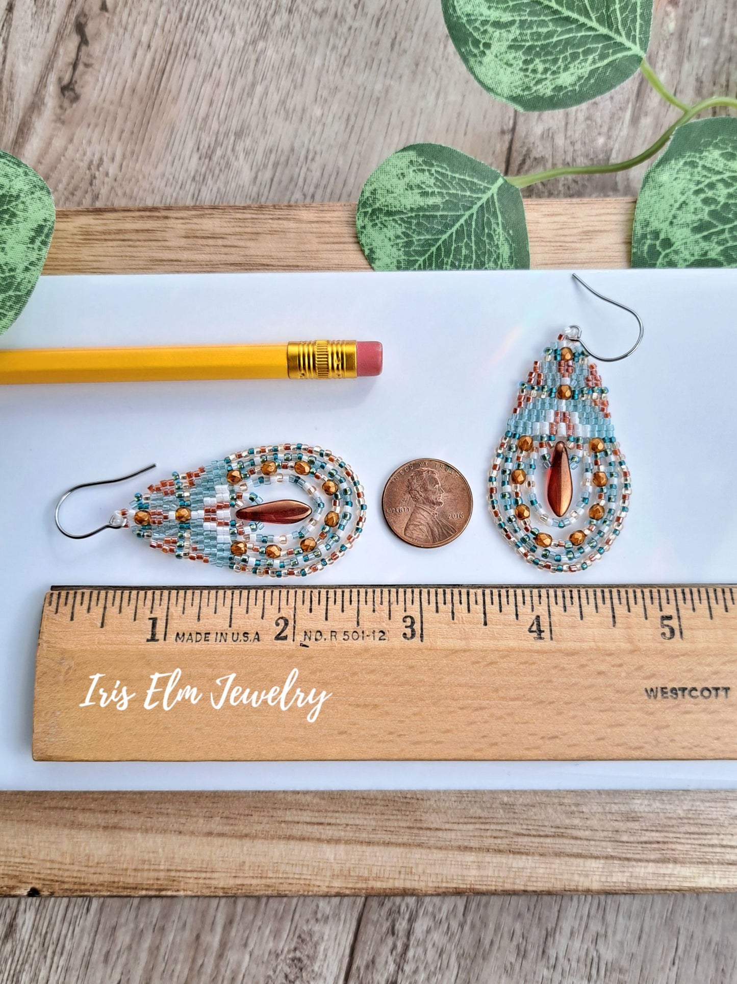 Two handmade 2.5 inch beaded teardrop dangle earrings displayed on a white surface with a wooden ruler, a pencil, a penny, and green leaves. The earrings feature light blue, white, and copper-orange beads with a central copper-red drop.