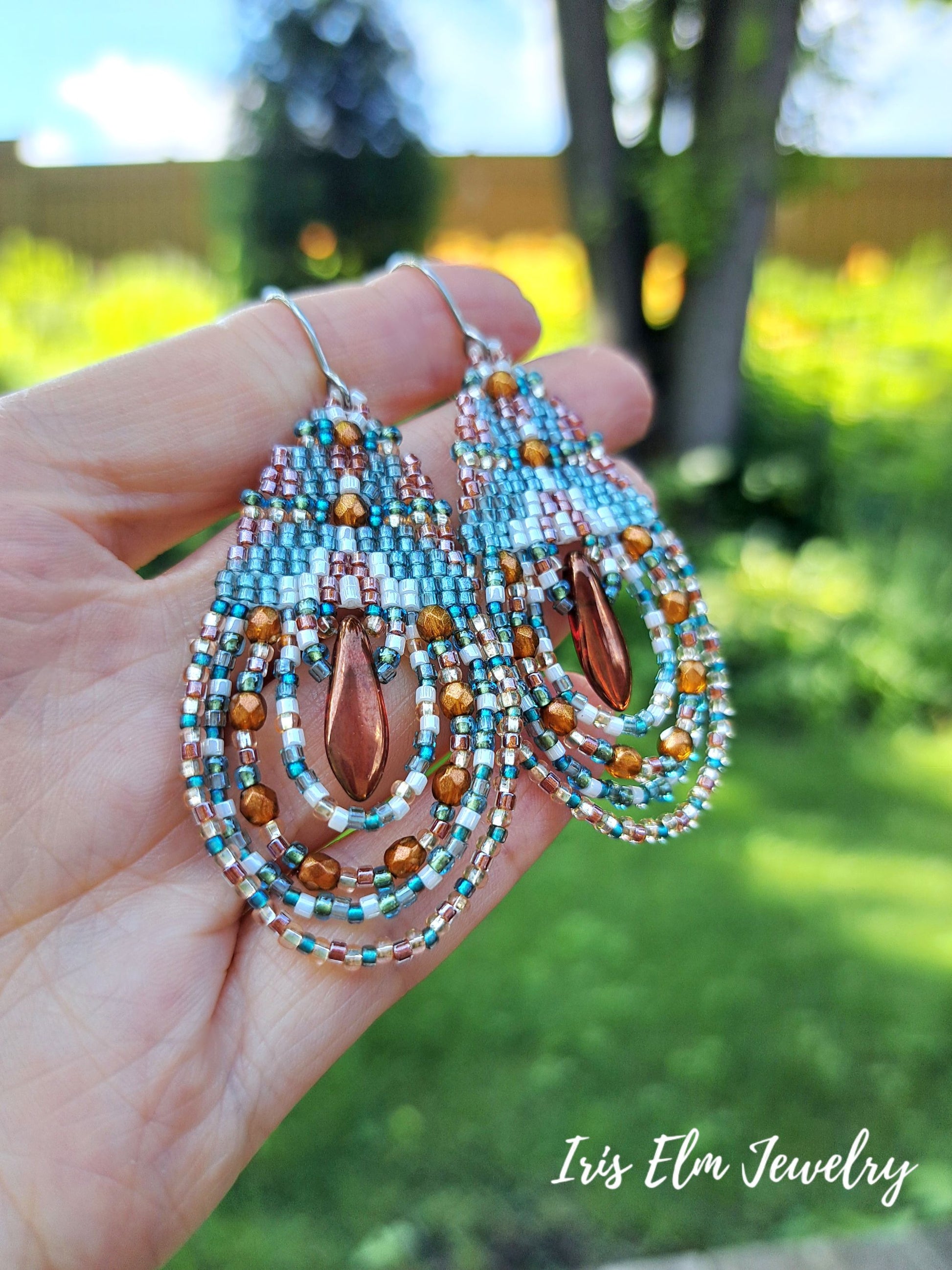 A hand holds a pair of large teardrop-shaped beaded earrings outdoors with a blurred green garden background. The earrings feature turquoise, white, and copper-colored seed beads with a central copper teardrop bead.