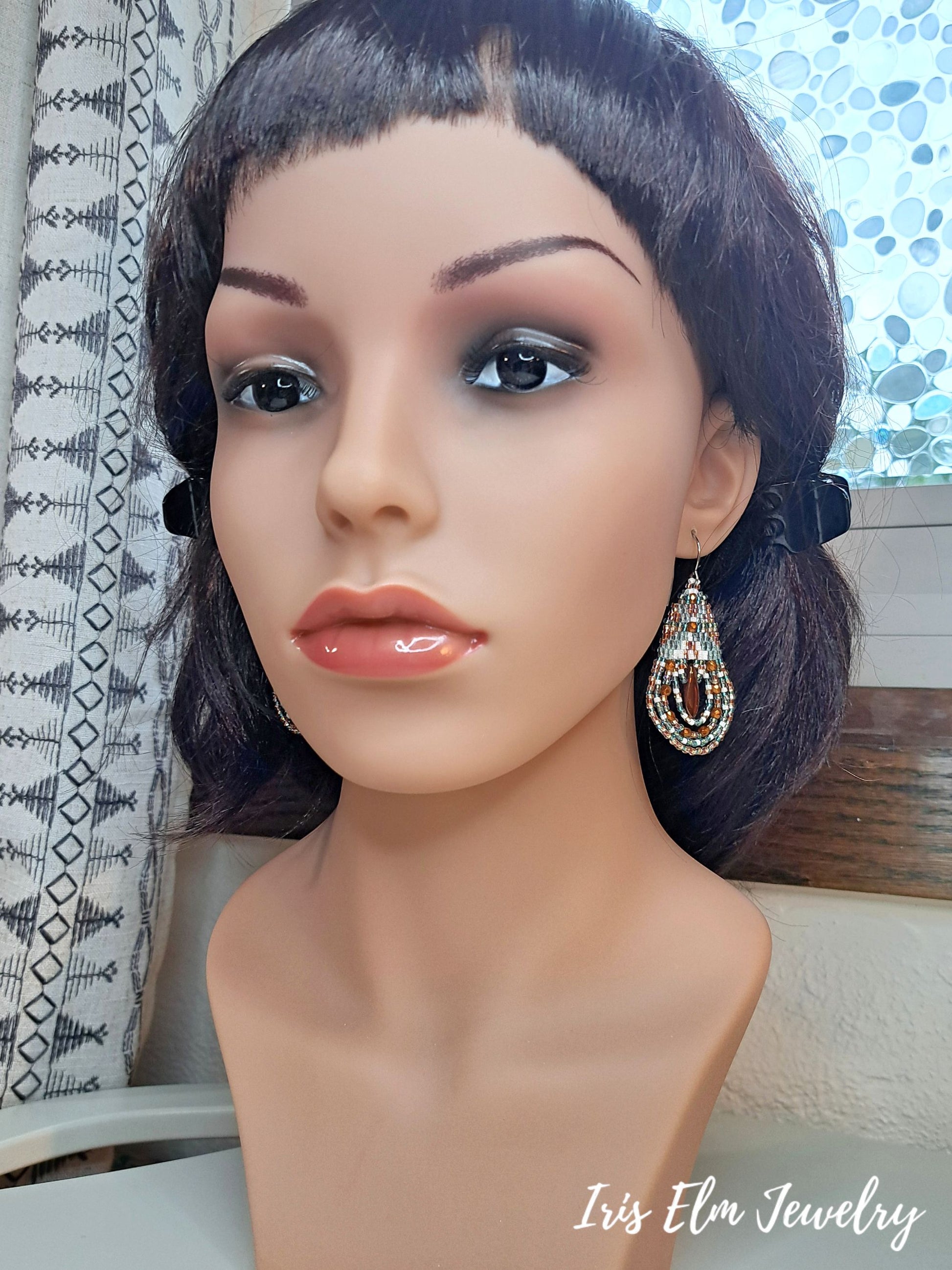 A mannequin bust wearing a pair of dangling teardrop earrings with burnt orange, turquoise, and white glass beads, set against an indoor background with a patterned curtain.
