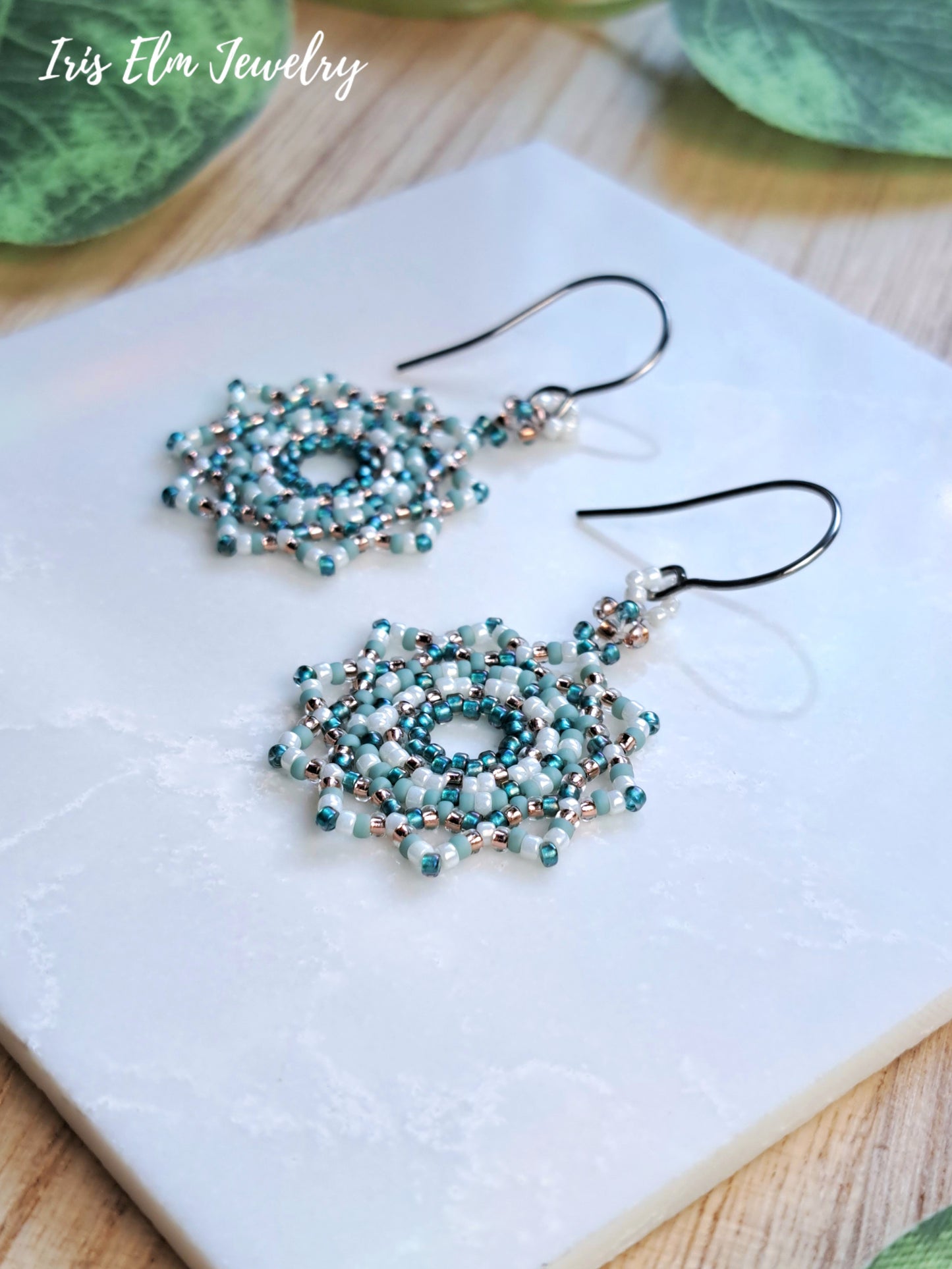 A pair of handmade beaded dangle earrings with intricate teal, white, and copper beadwork on steel fishhook wires, displayed on a white stone slab with green leaves in the background. The brand name 'Iris Elm Jewelry' is visible in the top left corner.