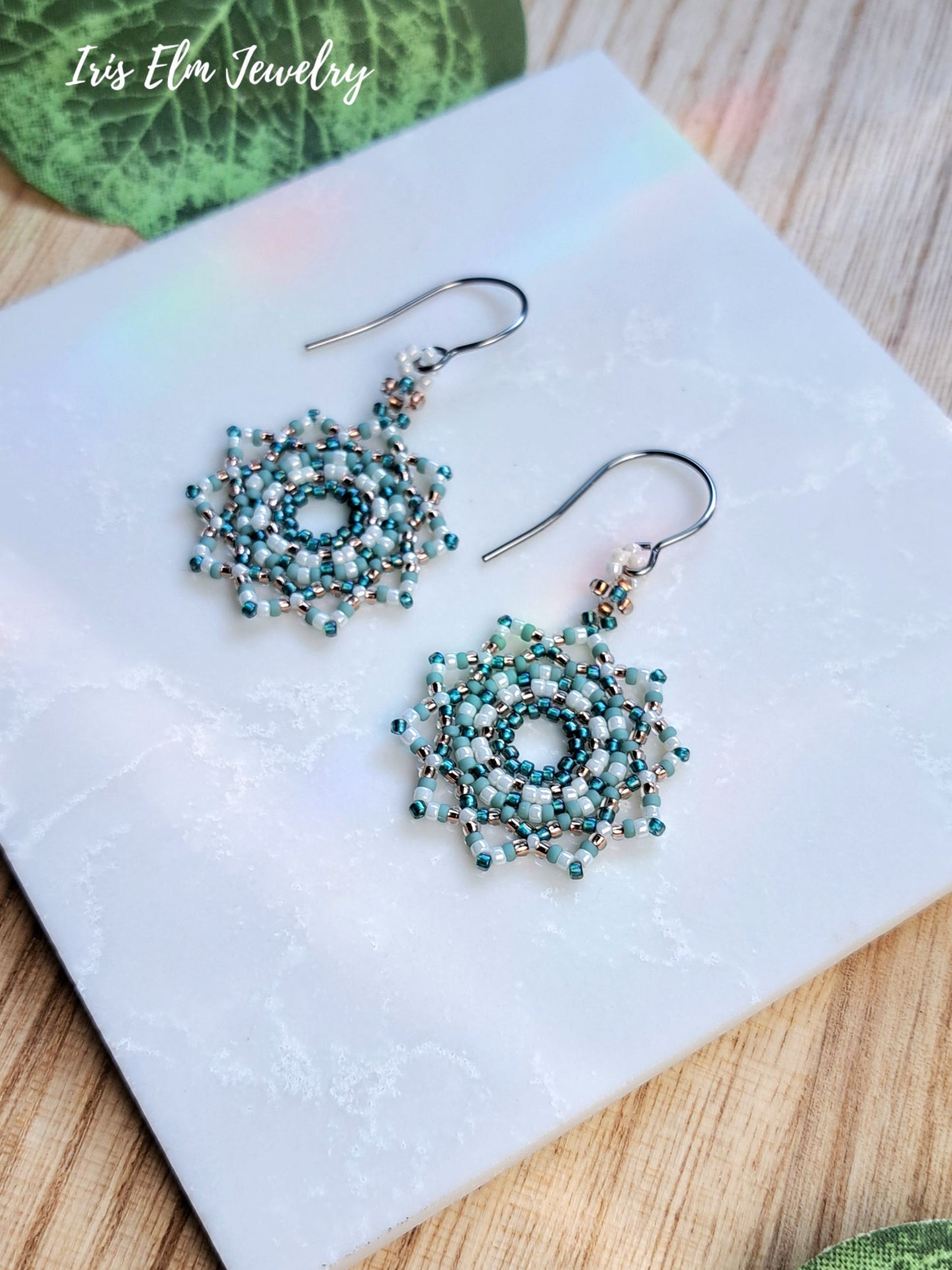 A pair of handmade beaded dangle mandala earrings with intricate circular patterns in teal, white, and copper-tone glass seed beads, displayed on a white marble tile.