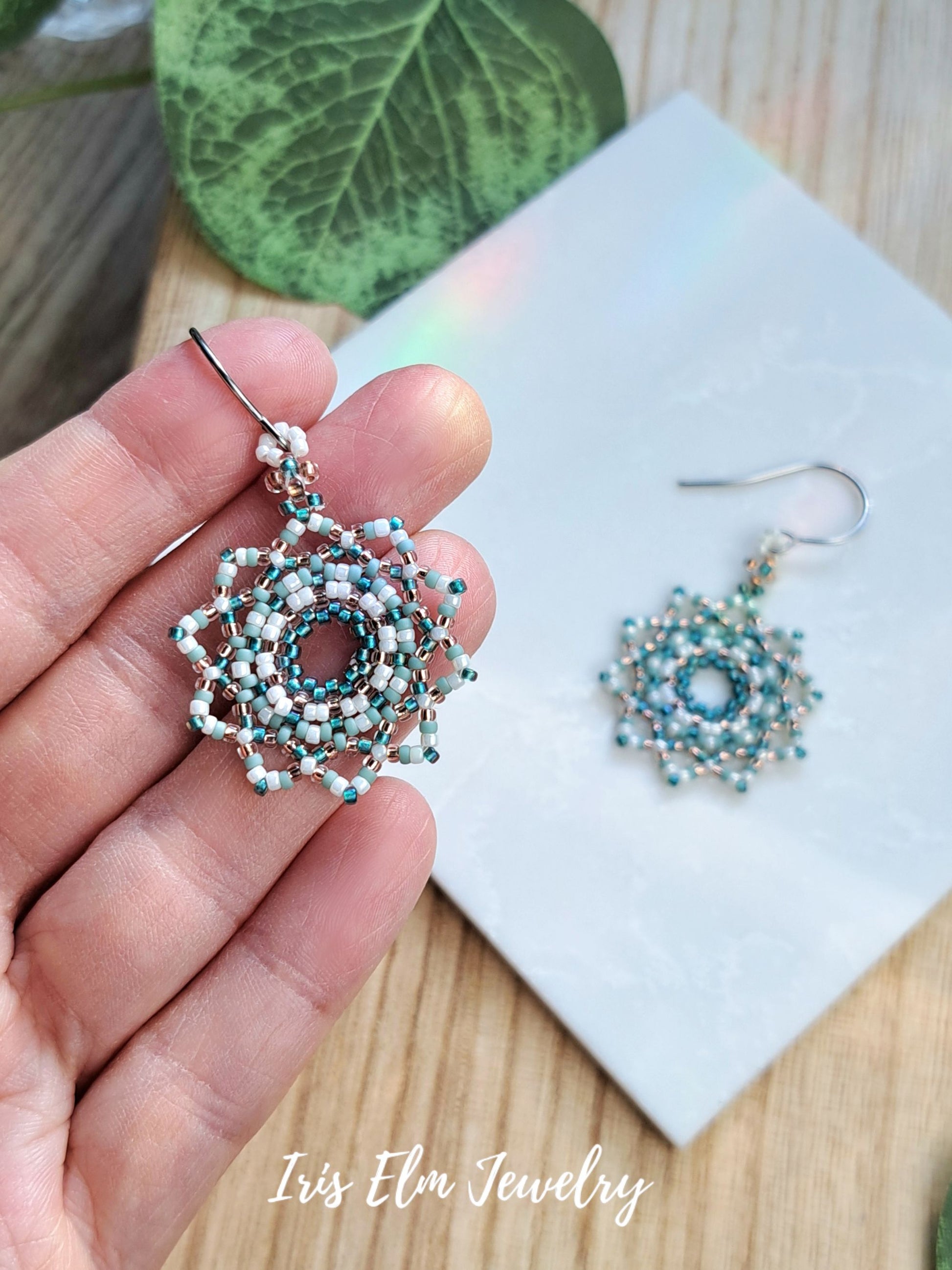 A hand holds a pair of intricate beaded dangle earrings with a silver-toned fishhook. The earrings feature a circular center surrounded by layered geometric patterns in light blue, teal, and white beads.