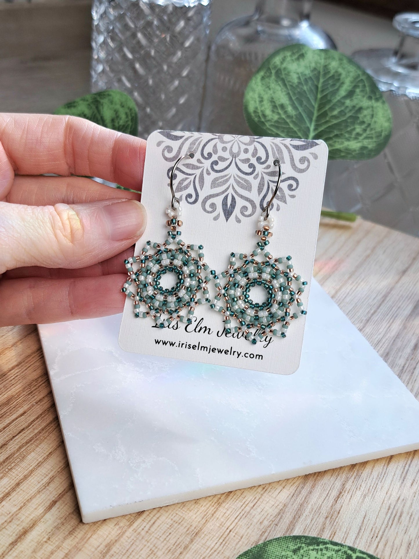 A hand holds a pair of intricate beaded dangle earrings on a display card. The earrings feature silver-toned metal frames and rows of small green and white beads.