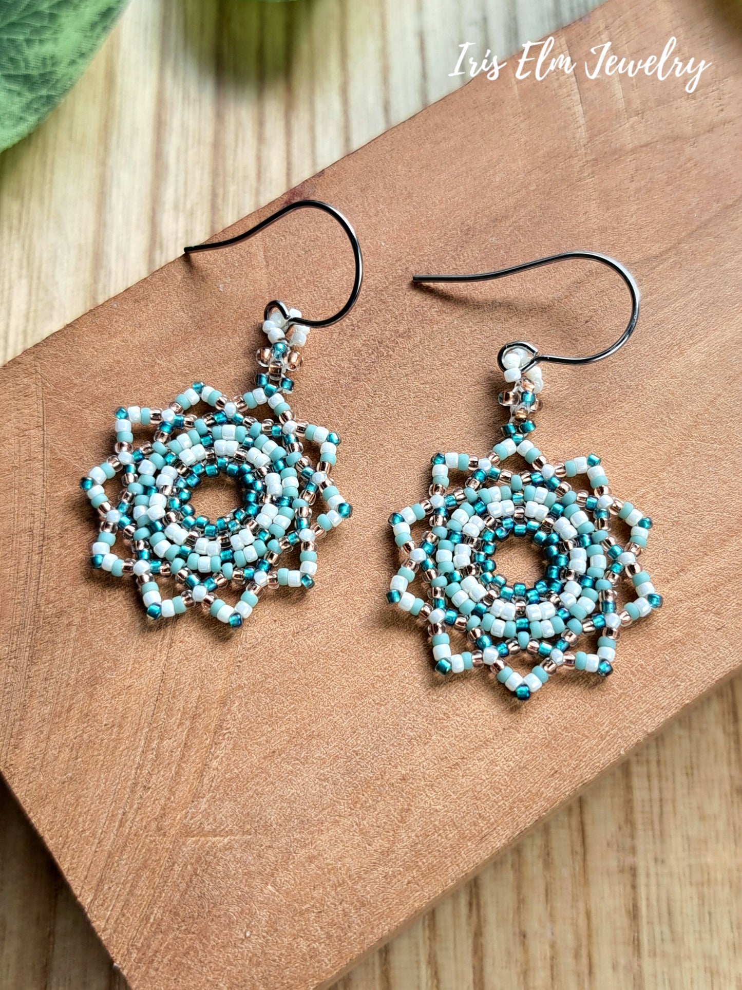 A pair of handmade beaded dangle earrings with steel fishhook wires, displayed on a brown wooden block. The earrings feature intricate circular mandala-like beadwork in turquoise, light blue, white, and copper.