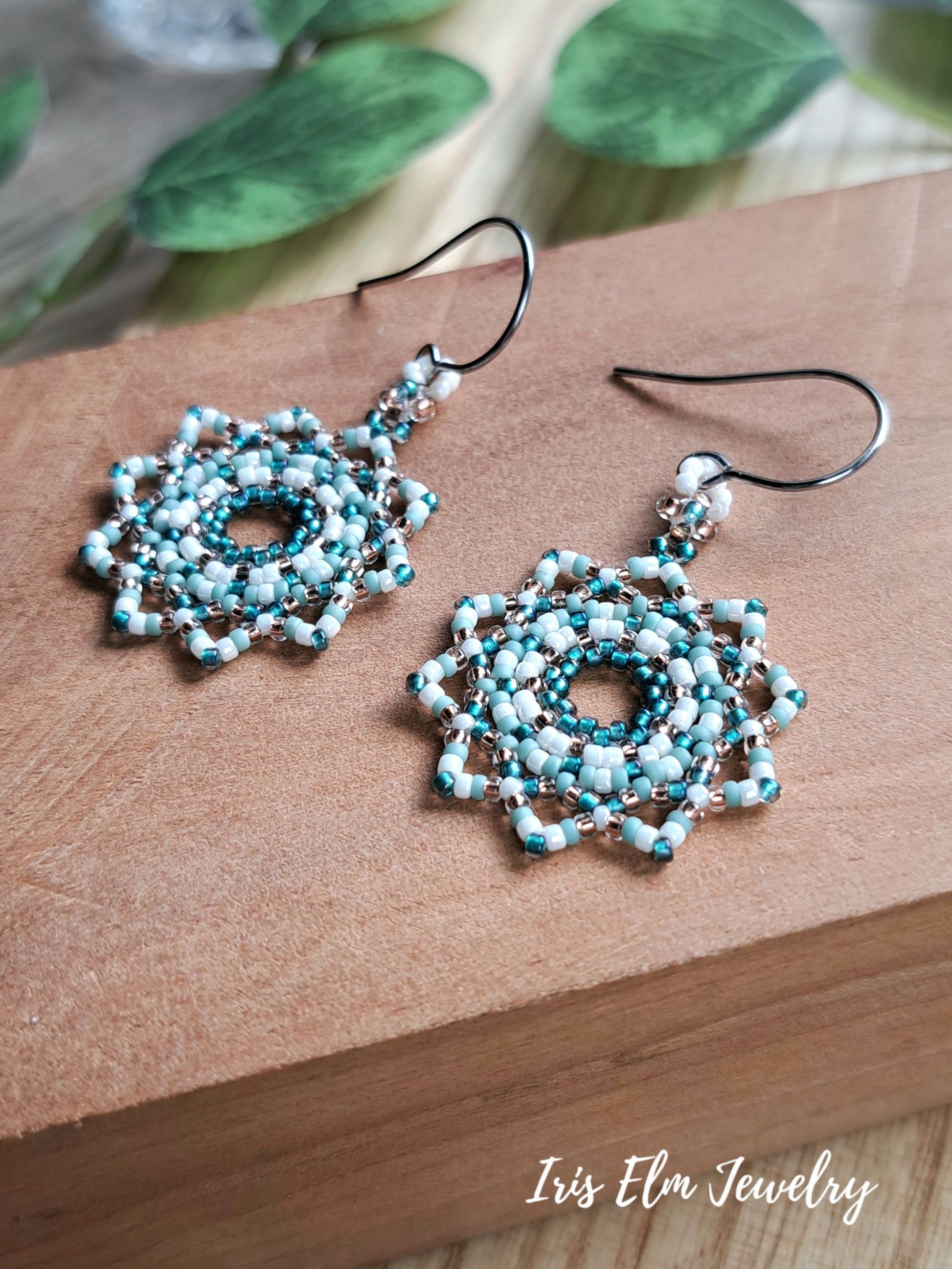 A pair of handmade beaded dangle earrings with intricate mandala circular patterns in light blue, white, and teal, suspended from steel fishhook wires. The earrings are displayed on a brown wooden block with blurred green leaves in the background.