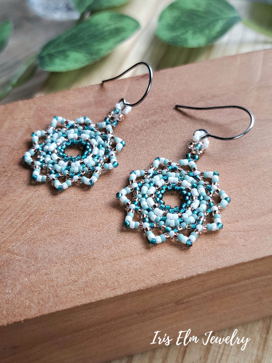 A pair of handmade beaded dangle earrings with intricate mandala circular patterns in light blue, white, and teal, suspended from steel fishhook wires. The earrings are displayed on a brown wooden block with blurred green leaves in the background.
