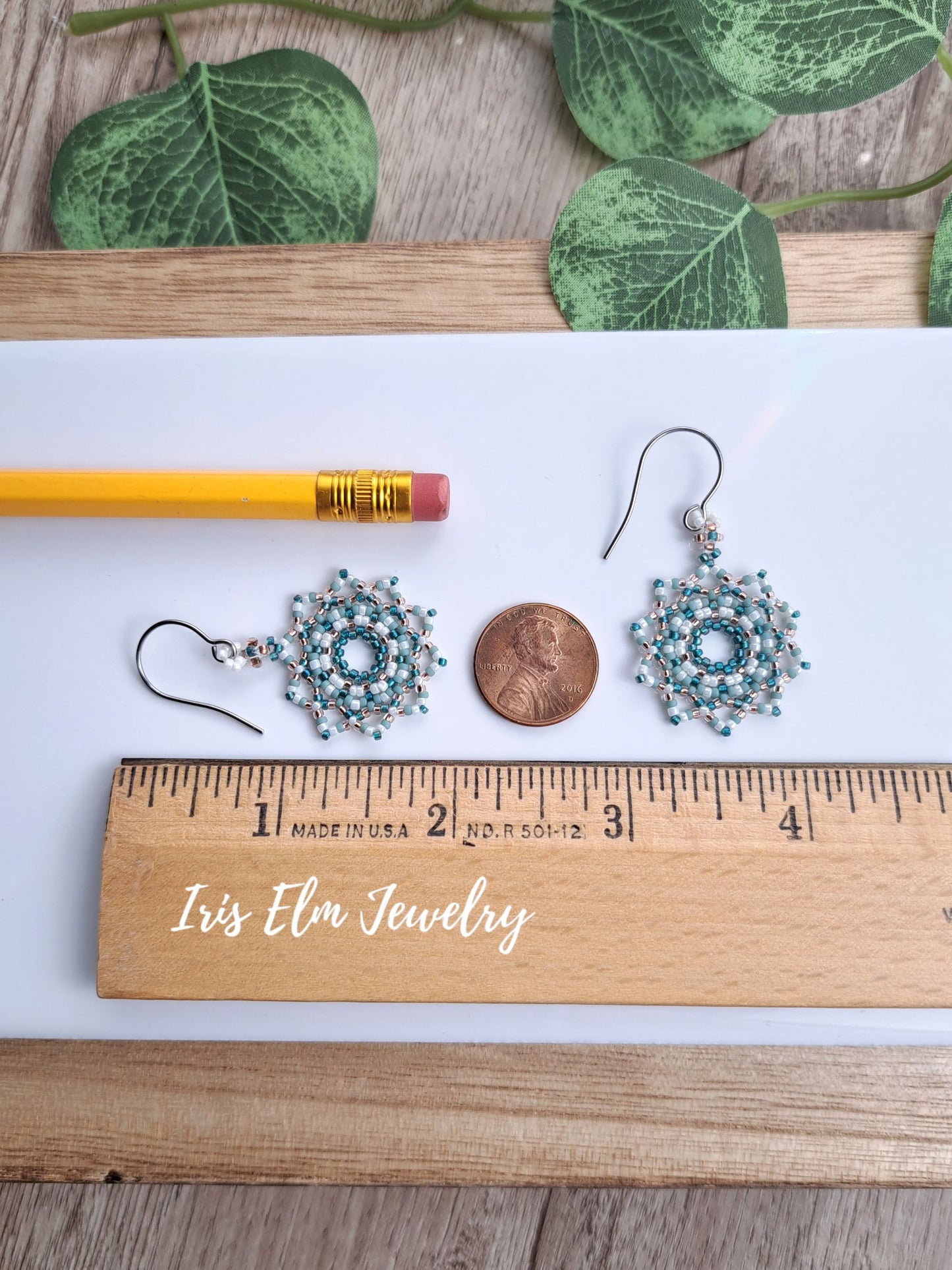 A pair of 1-7/8 inch long handmade beaded dangle earrings with turquoise and silver beads, displayed on a white sheet of paper next to a yellow pencil, a penny for scale, and a wooden ruler. The text 'Iris Elm Jewelry' is written on the ruler.