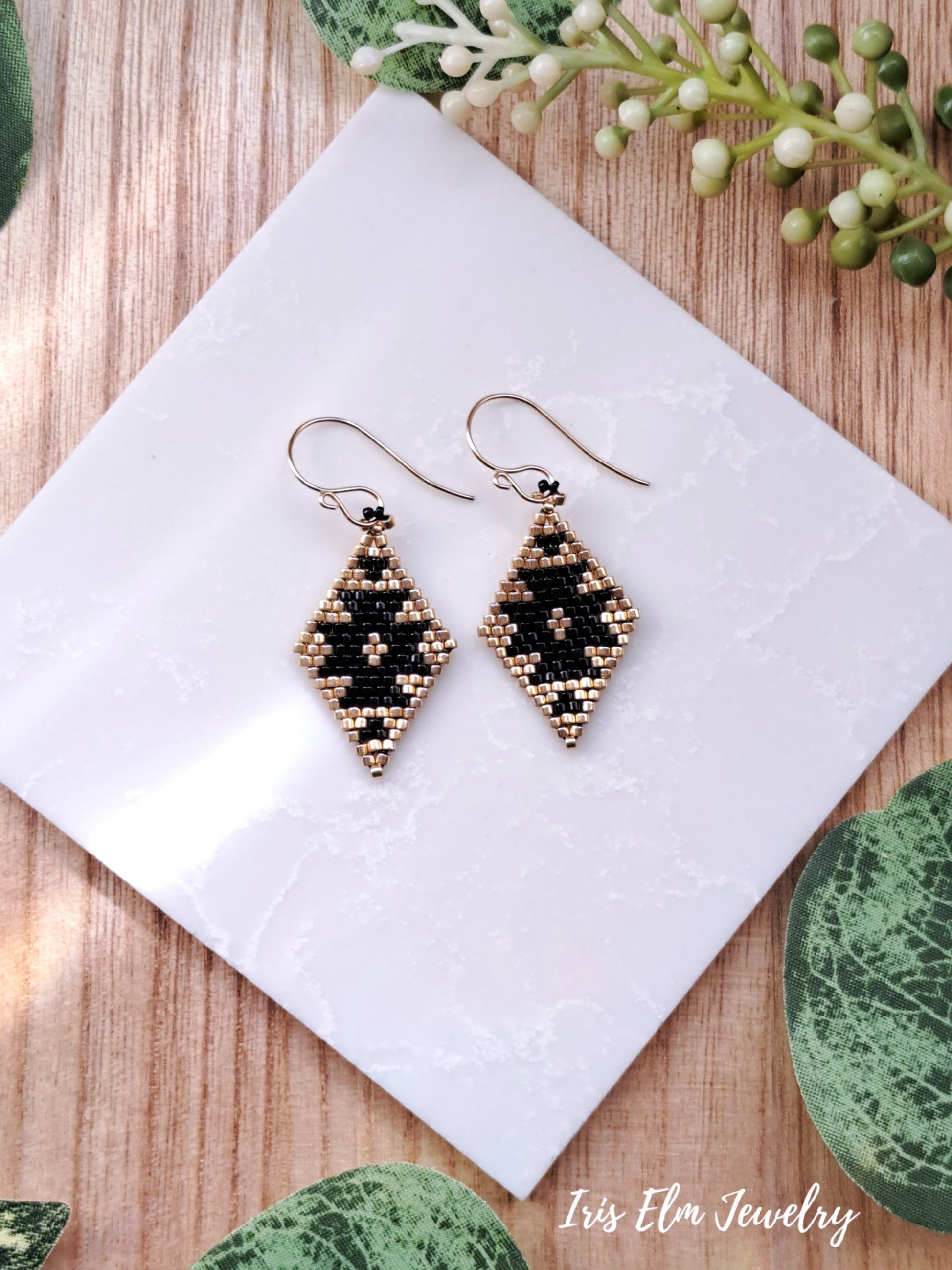 Pair of beaded black and gold southwestern seed bead diamond shape earrings on a white tile with greenery in the background