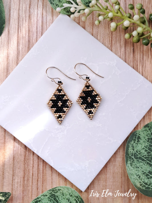 Pair of beaded black and gold southwestern seed bead diamond shape earrings on a white tile with greenery in the background