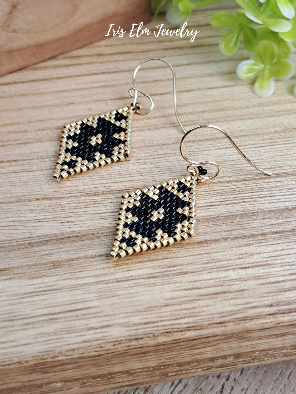 A pair of handmade beaded dangle earrings with geometric black and gold seed bead patterns on gold-tone fishhook wires, displayed on a wooden surface with a small green plant in the background. The text 'Iris Elm Jewelry' is visible at the top.