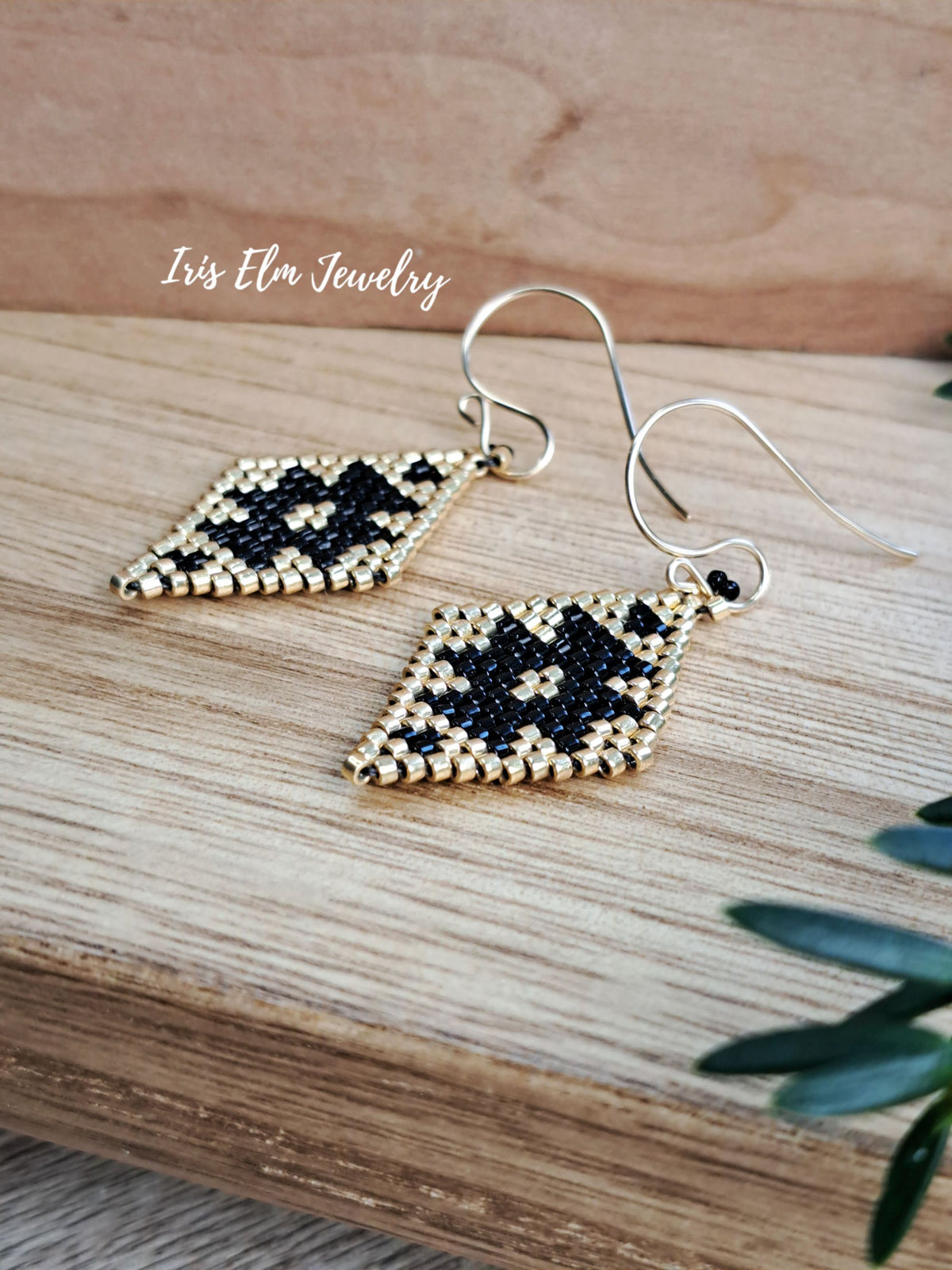 A pair of handmade beaded dangle earrings with a geometric diamond pattern in black and gold, hanging from goldfilled fishhook wires. The earrings are displayed on a light wood surface with a wooden background, with the brand name 'Iris Elm Jewelry' written in white script.