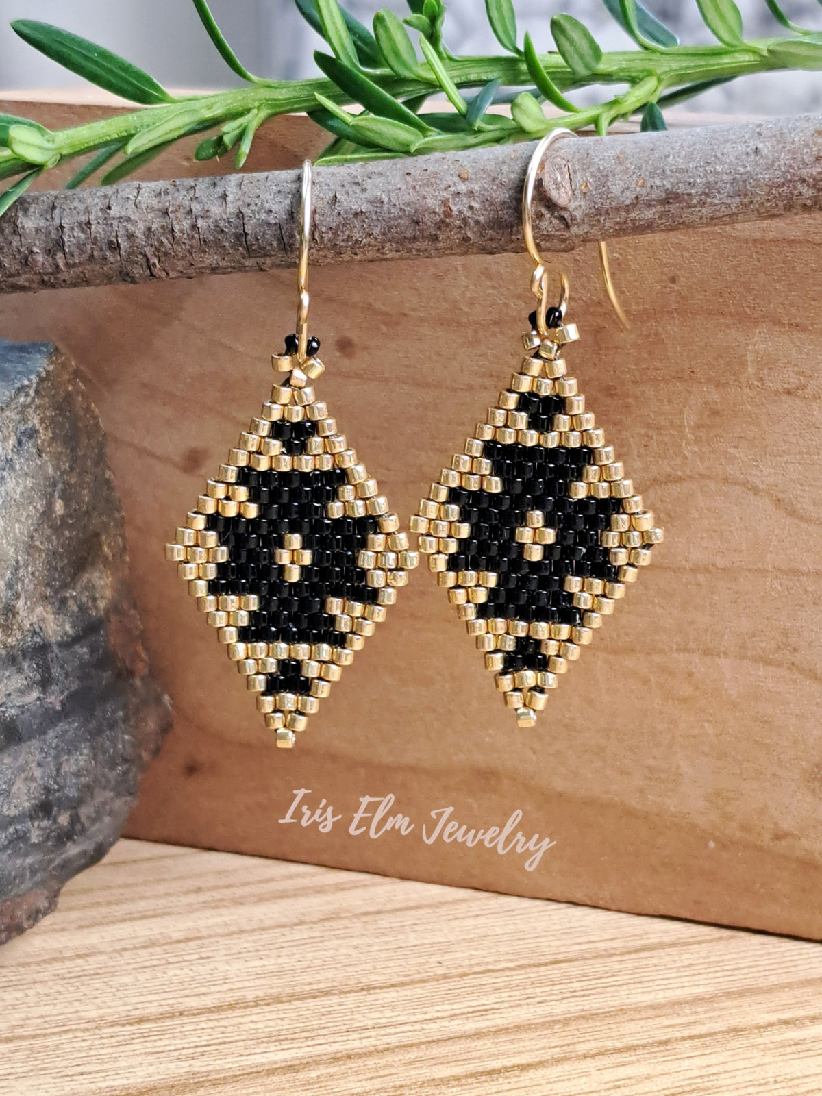 A pair of handmade beaded dangle earrings with a geometric diamond pattern in black and gold seed beads, hanging from gold fishhook wires. The earrings are displayed on a rustic wooden block with a green sprig and a rock prop, with the brand name 'Iris Elm Jewelry' printed on the wood.