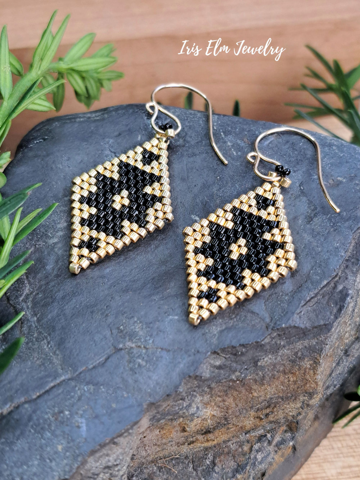 A pair of handmade beaded dangle earrings with a geometric black, gold, pattern, displayed on a dark slate rock with green foliage. The brand name 'Iris Elm Jewelry' is visible in the top right corner.