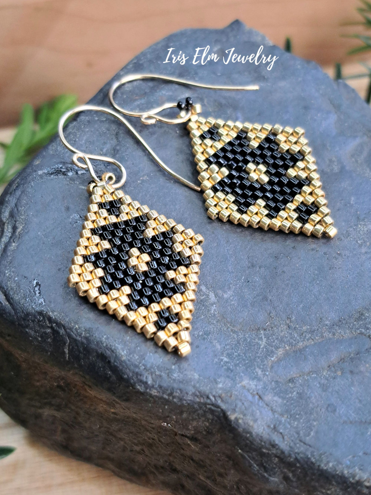 A pair of handmade beaded dangle earrings with geometric black and gold patterns on diamond-shaped charms, displayed on a dark slate stone. The earrings feature gold-toned metal fishhook wires.