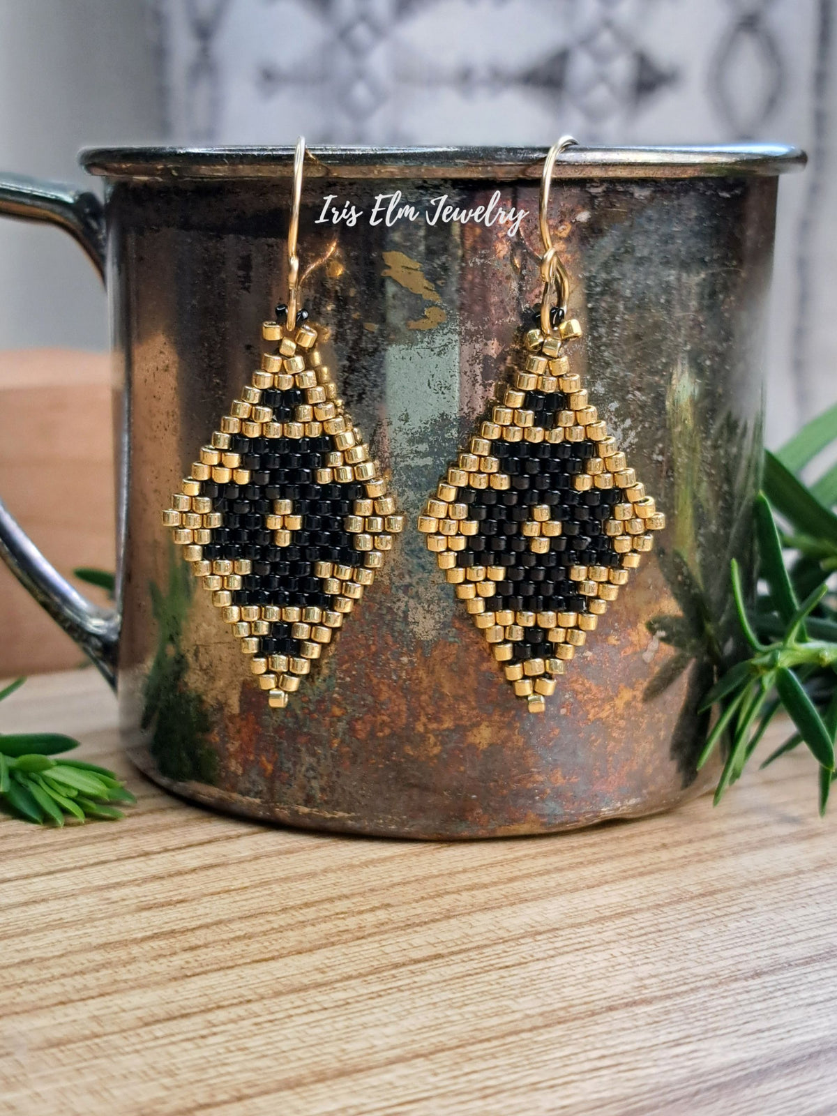 A pair of handmade beaded dangle earrings with a black and gold geometric diamond pattern, hanging from gold-toned hooks. They are displayed on the rim of a rustic, distressed copper mug, set on a wooden surface with green foliage in the background.