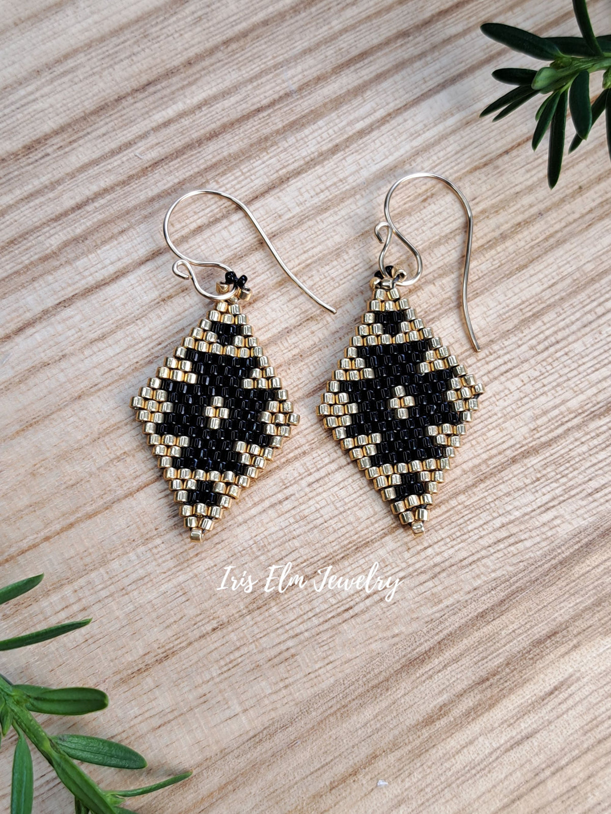 A pair of handmade beaded dangle earrings with a black and gold geometric pattern, hanging from goldfilled fishhook wires on a light wood background with green leaves in the corners.